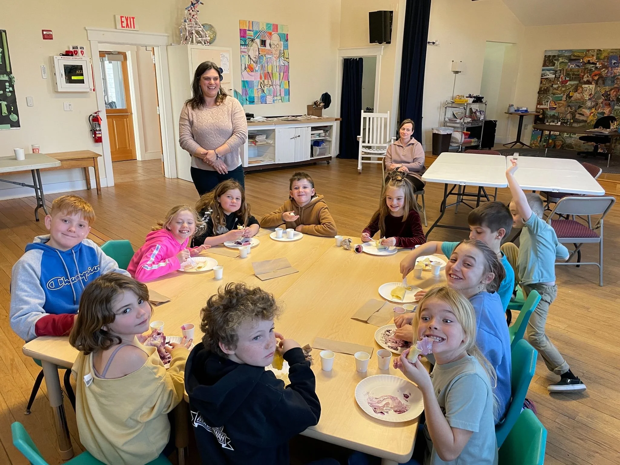 Children and adults gathered around a table during a birthday celebration in a community room, with some eating cake and others smiling at the camera.