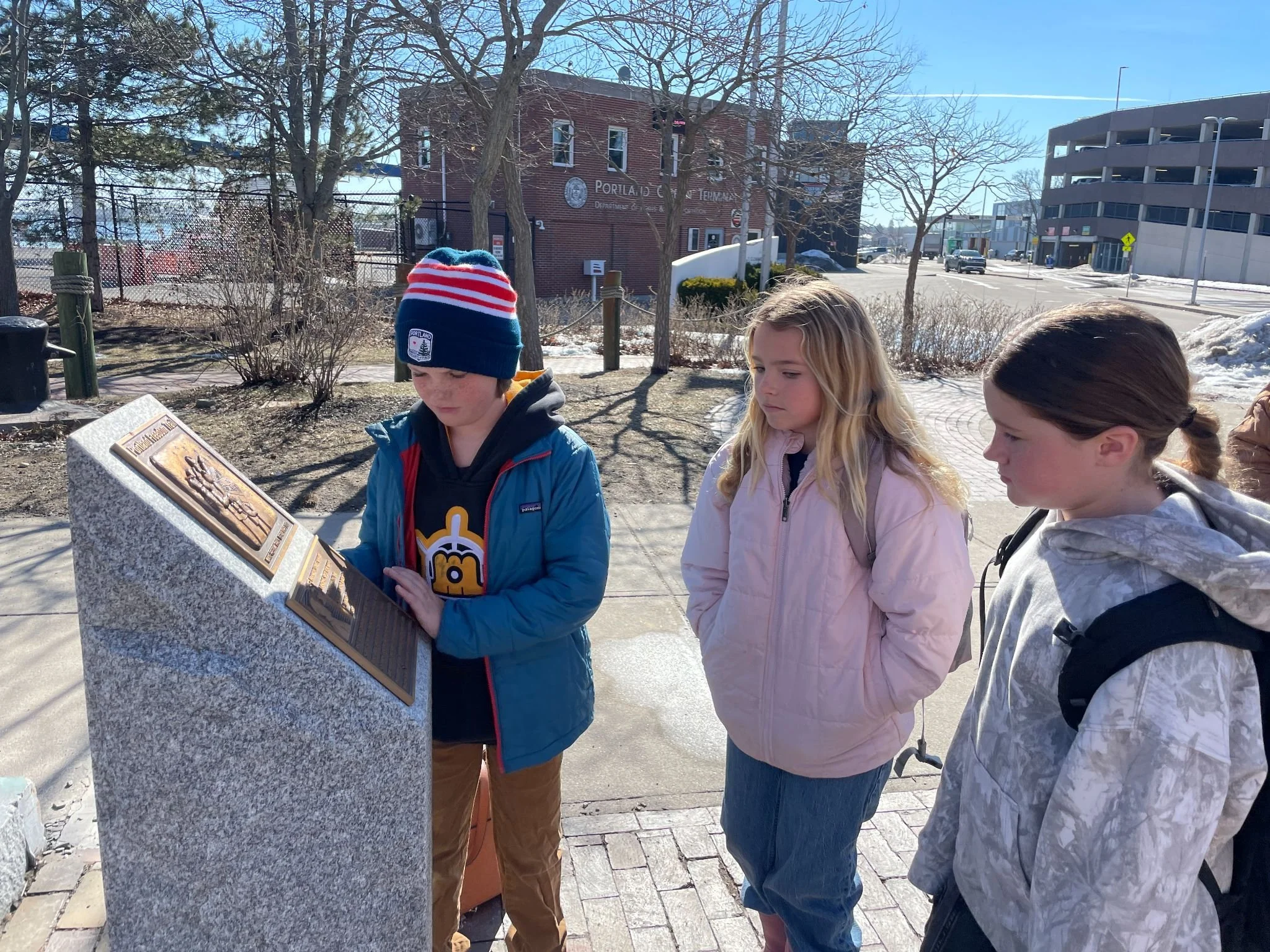 Four children standing outdoors, looking at a historical plaque mounted on a stone pedestal, with trees and buildings in the background on a sunny day.