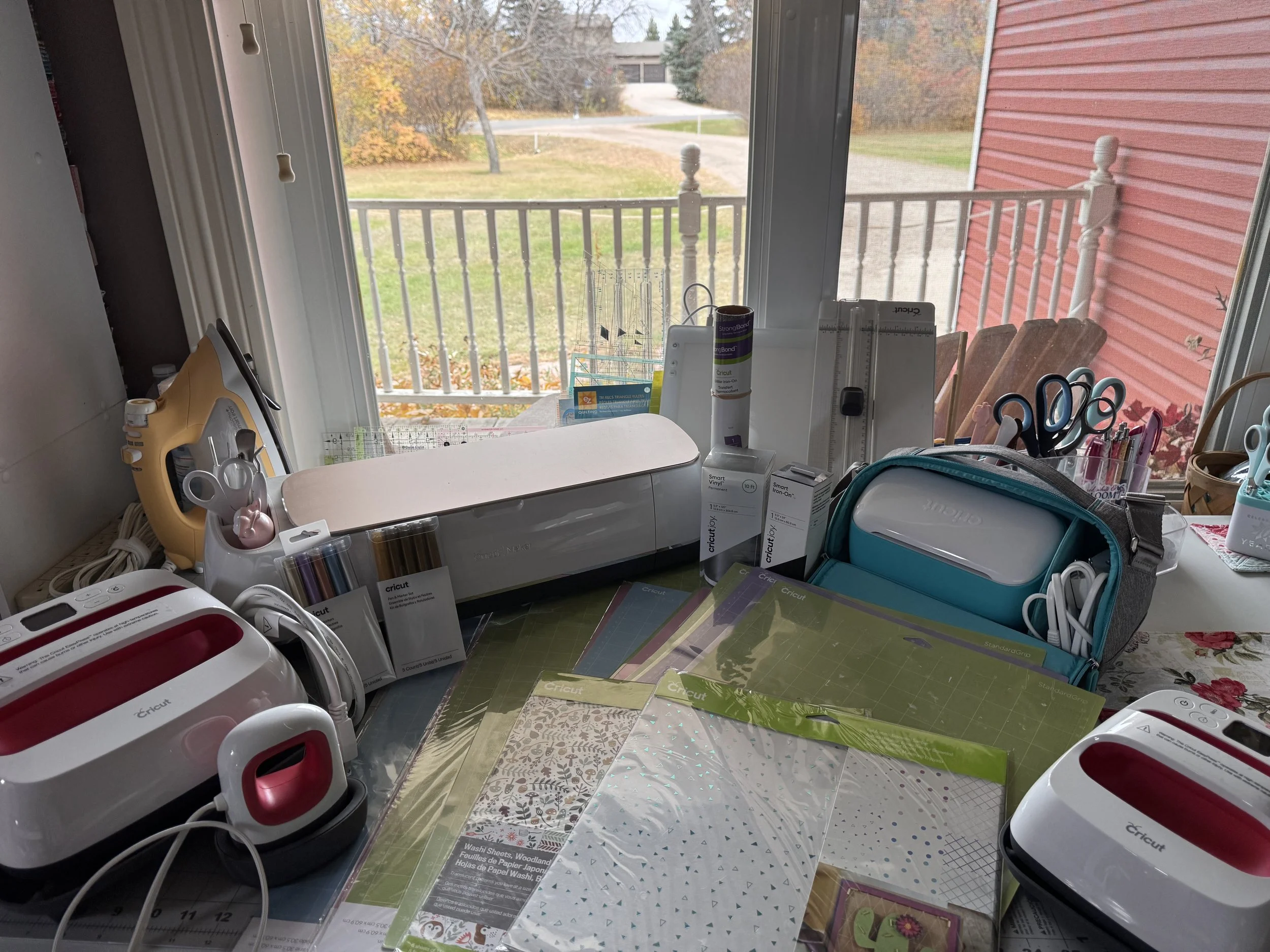 Cricut crafting workspace with cutting machines, heat presses, vinyl sheets, tools, and accessories beside a window.