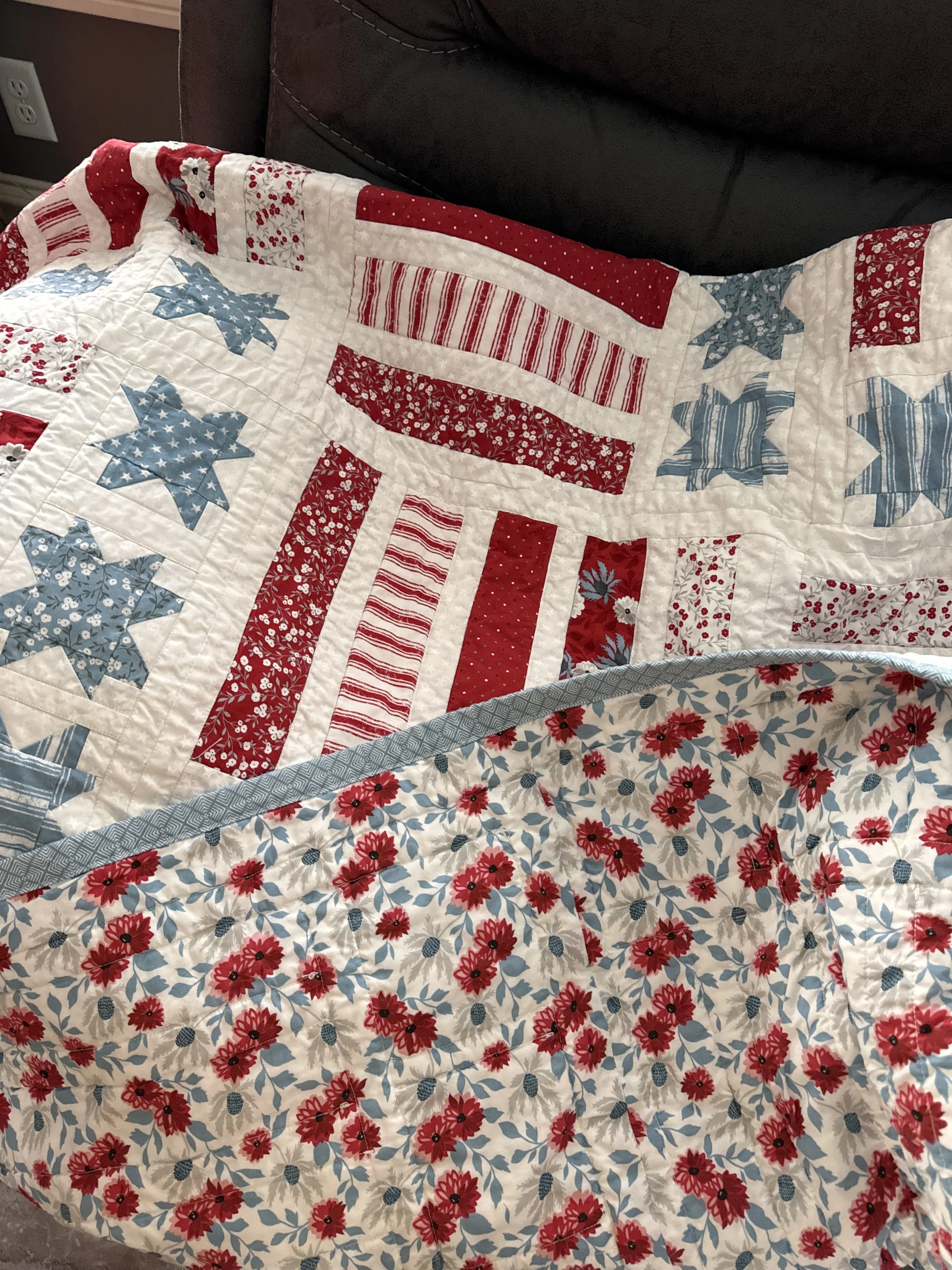 A quilt with a pattern of red, white, and blue stars and stripes in a patriotic design, draped over a bed with a floral sheet underneath.