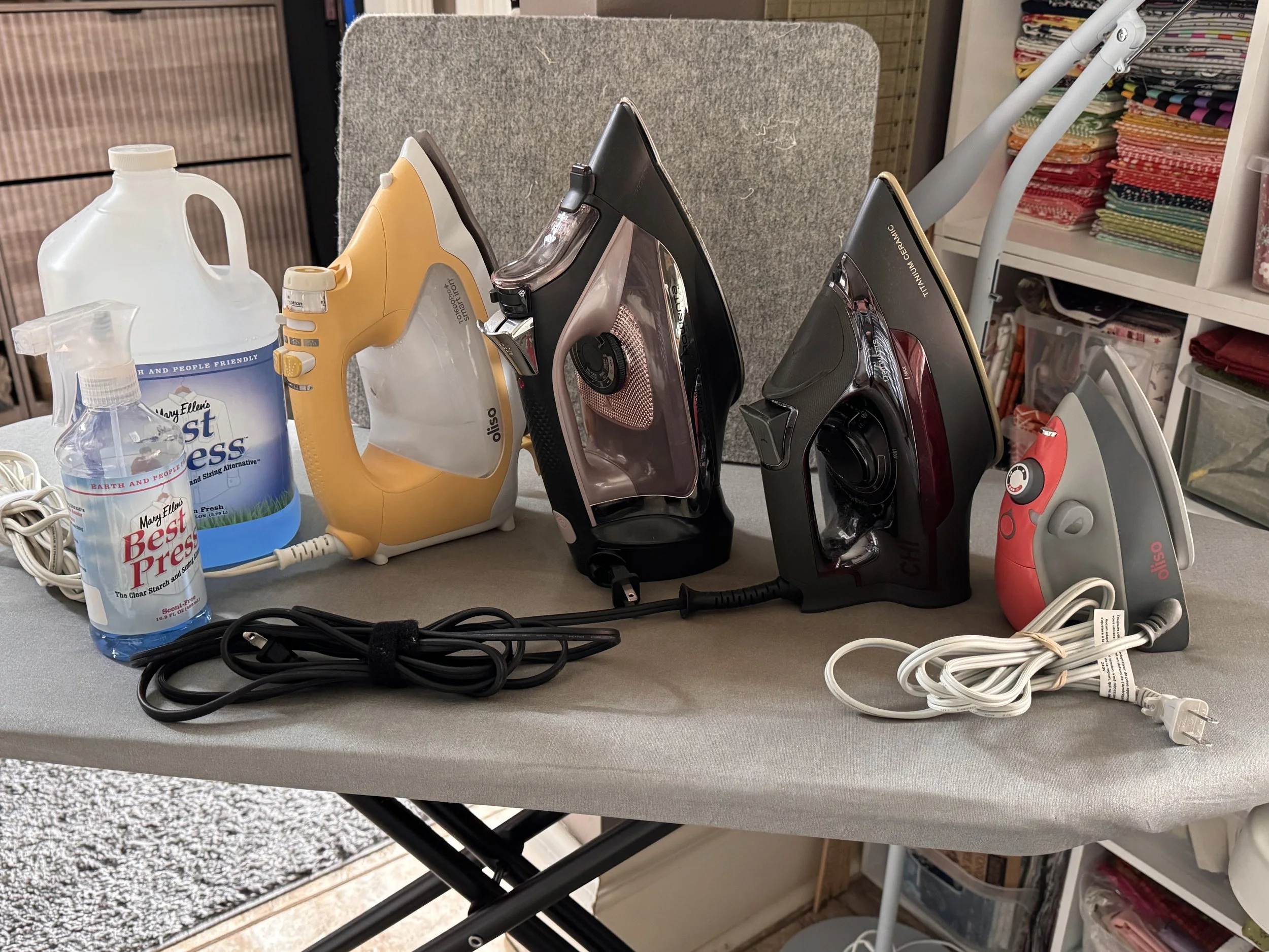 Assortment of household irons, cleaning spray bottles, and power cords on an ironing board in a sewing space