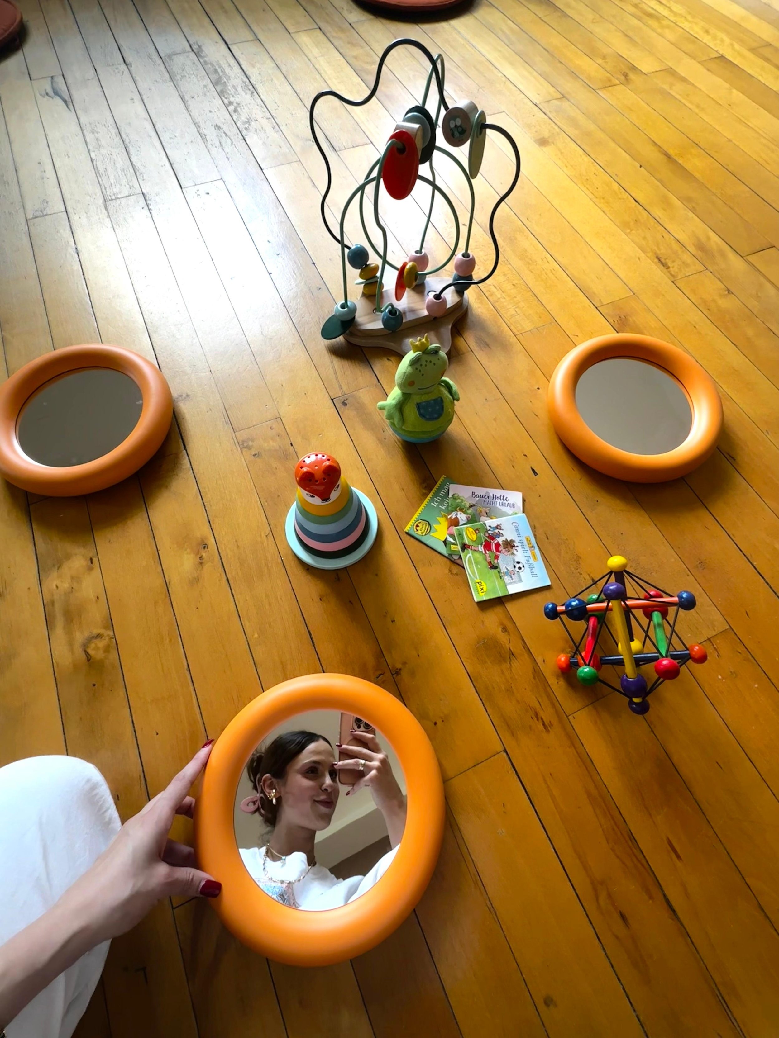 A woman taking a selfie in a mirror on a wooden floor surrounded by children's toys, including a bead maze, a stacking toy, and books.