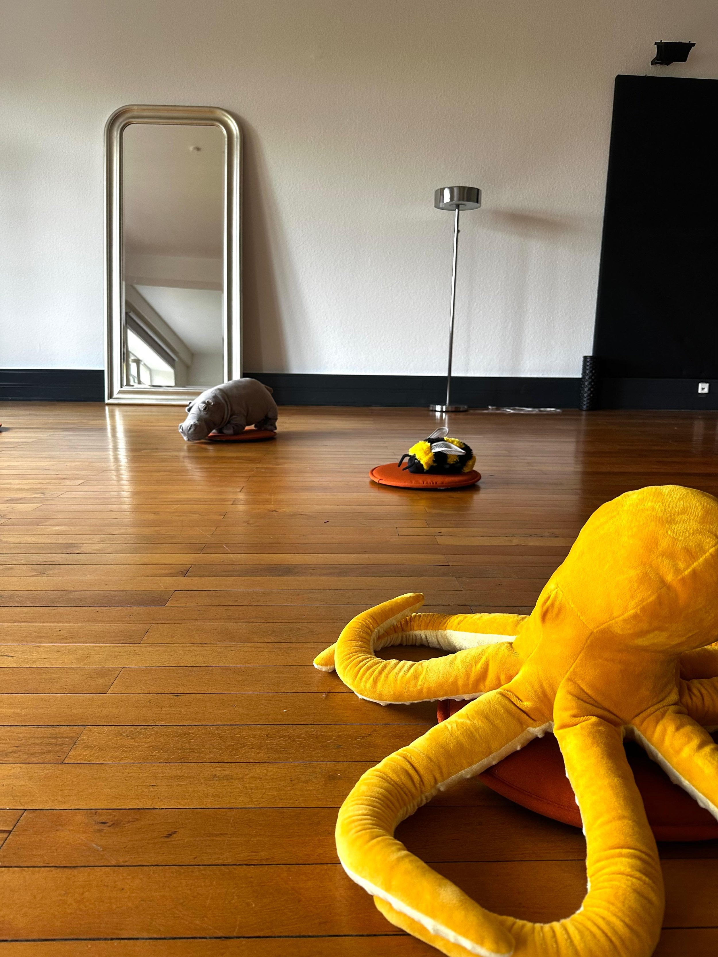 A yellow plush octopus toy with multiple legs is lying on a wooden floor near a black couch. In the background, a tall silver mirror, a modern silver floor lamp, and part of a black speaker or decoration are visible, with neutral-colored walls.