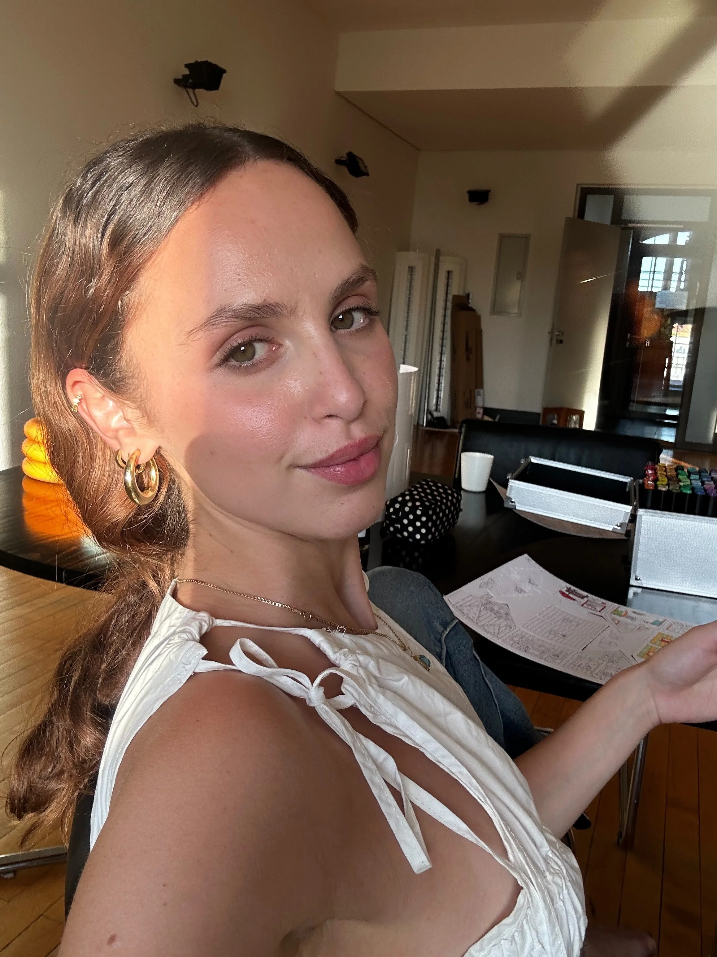 A woman with ash brown hair pinned back, sitting indoors and looking at the camera. There are papers and an open container on the table in front of her, with a background of a domestic space with some furniture and a glass door.