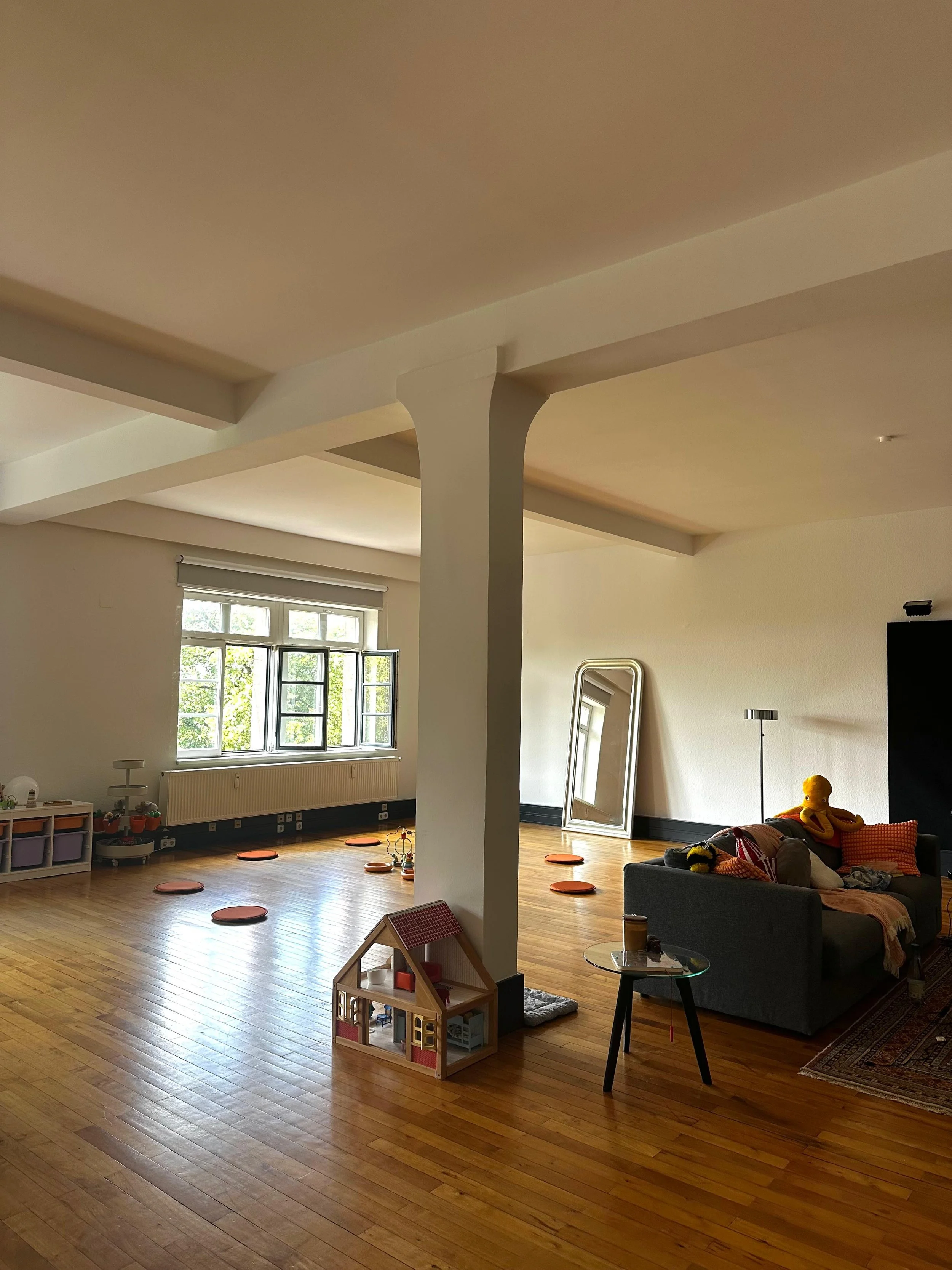 Living room with wooden flooring, large window, gray sofa with stuffed animals and pillows, a mirror leaning against the wall, and a small children's playhouse.
