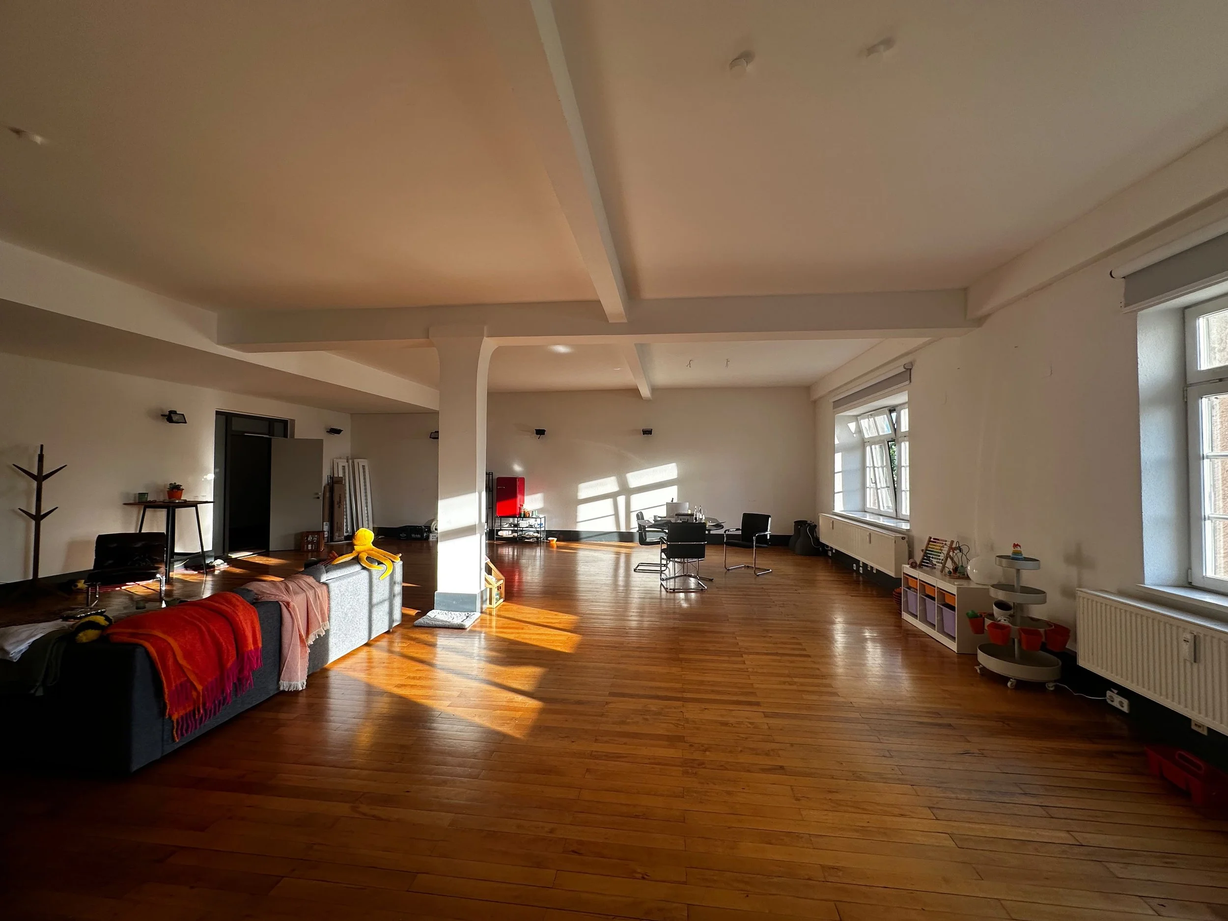 Spacious room with wooden floors, large windows with blinds, and a high ceiling with decorative beams. Contains furniture including a sofa with colorful blankets, a small table, black chairs, and children's toys.