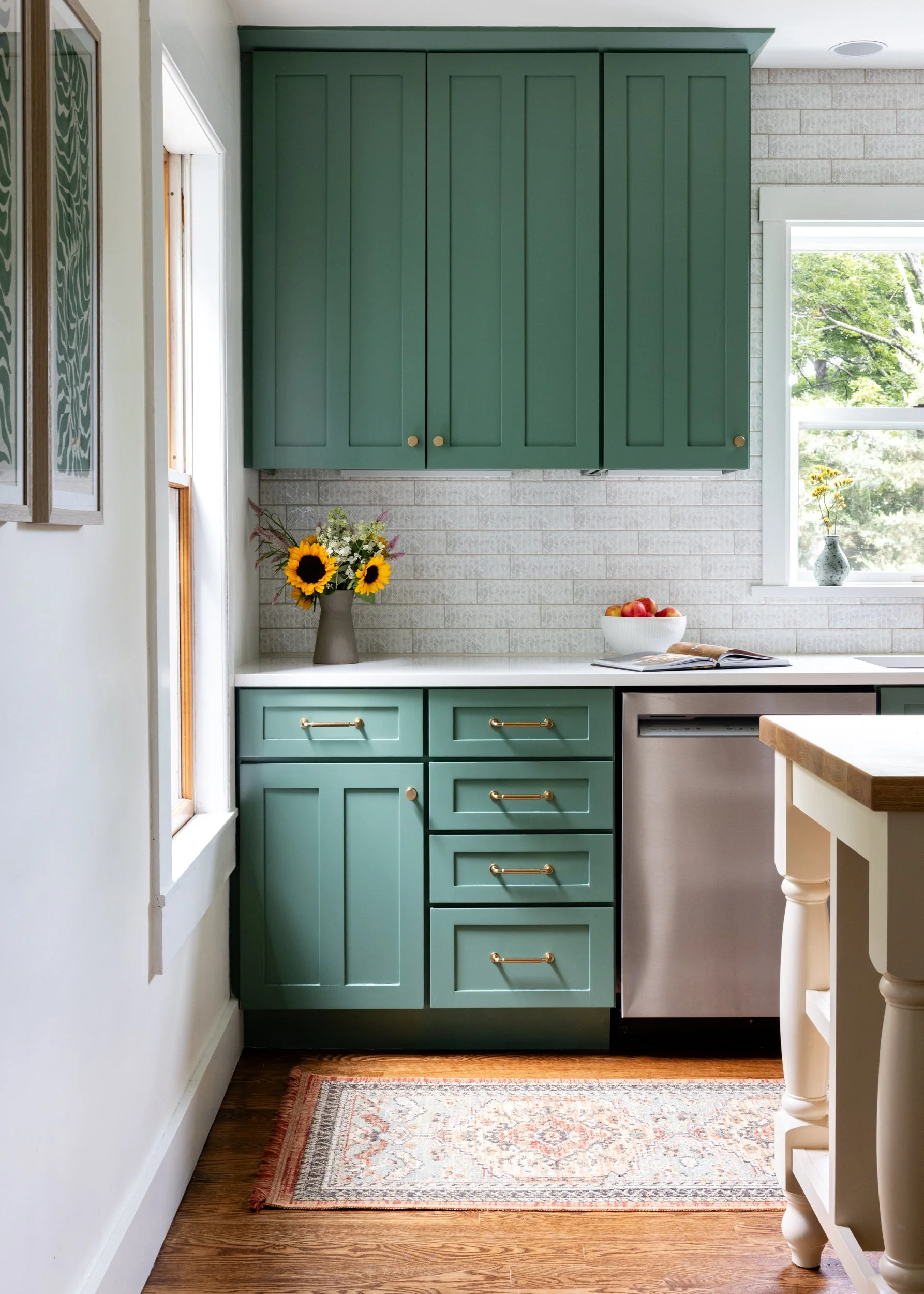 Kitchen with green cabinets, a vase of sunflowers, a bowl of apples, and an open book on the white countertop. There is a window showing greenery outside and a decorative rug on the wooden floor.