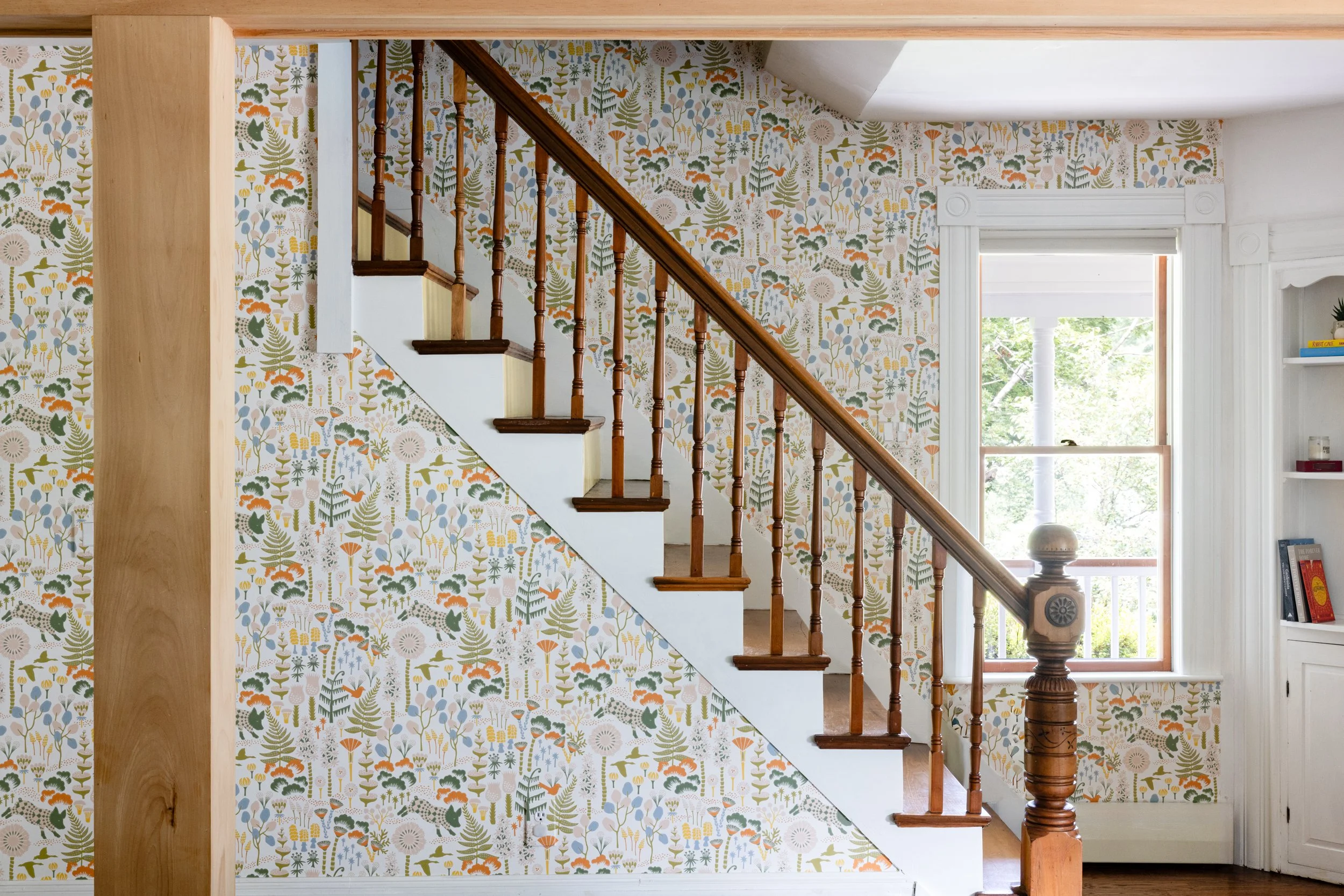 Interior view of a staircase with wooden banister next to a wallpapered wall with a colorful floral and animal pattern. A window and built-in bookshelf are visible.