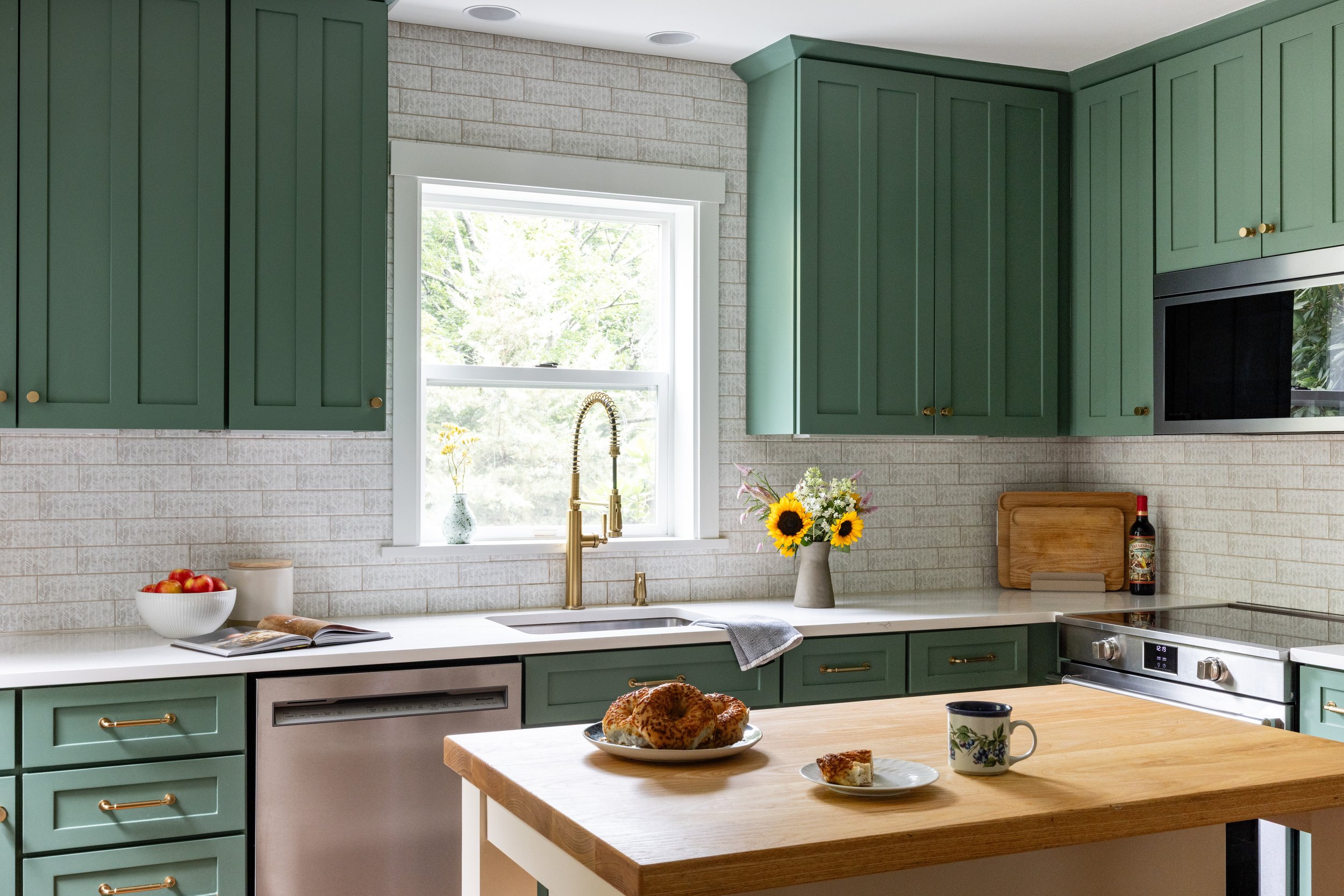 A cozy kitchen with green cabinets, white countertops, a window with a white frame, a vase with sunflowers, a bowl of apples, a loaf of bread, a mug, and a plate with a piece of cake.