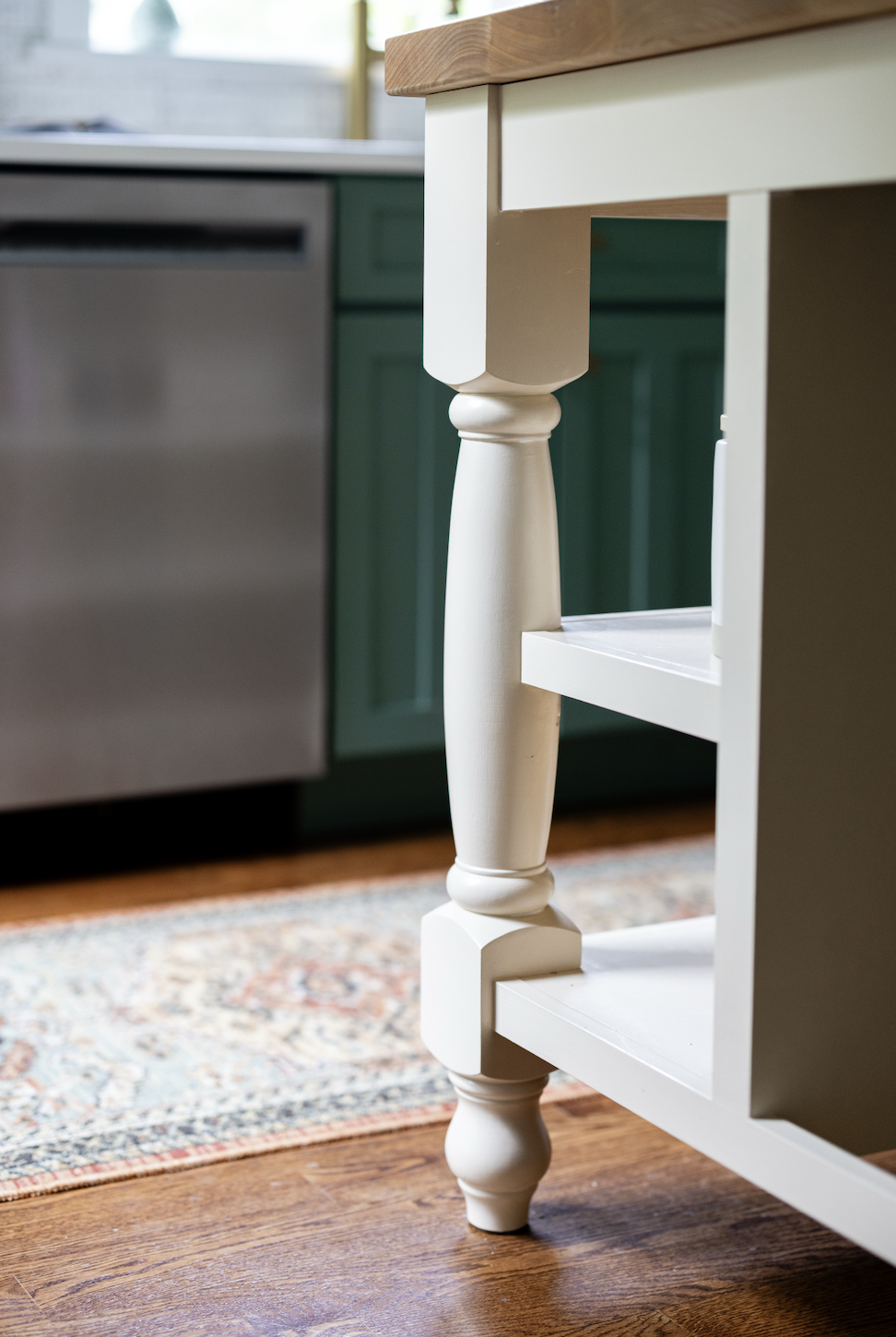 Close-up of a kitchen island with white legs, a wooden top, and a background of green and stainless steel kitchen cabinets.