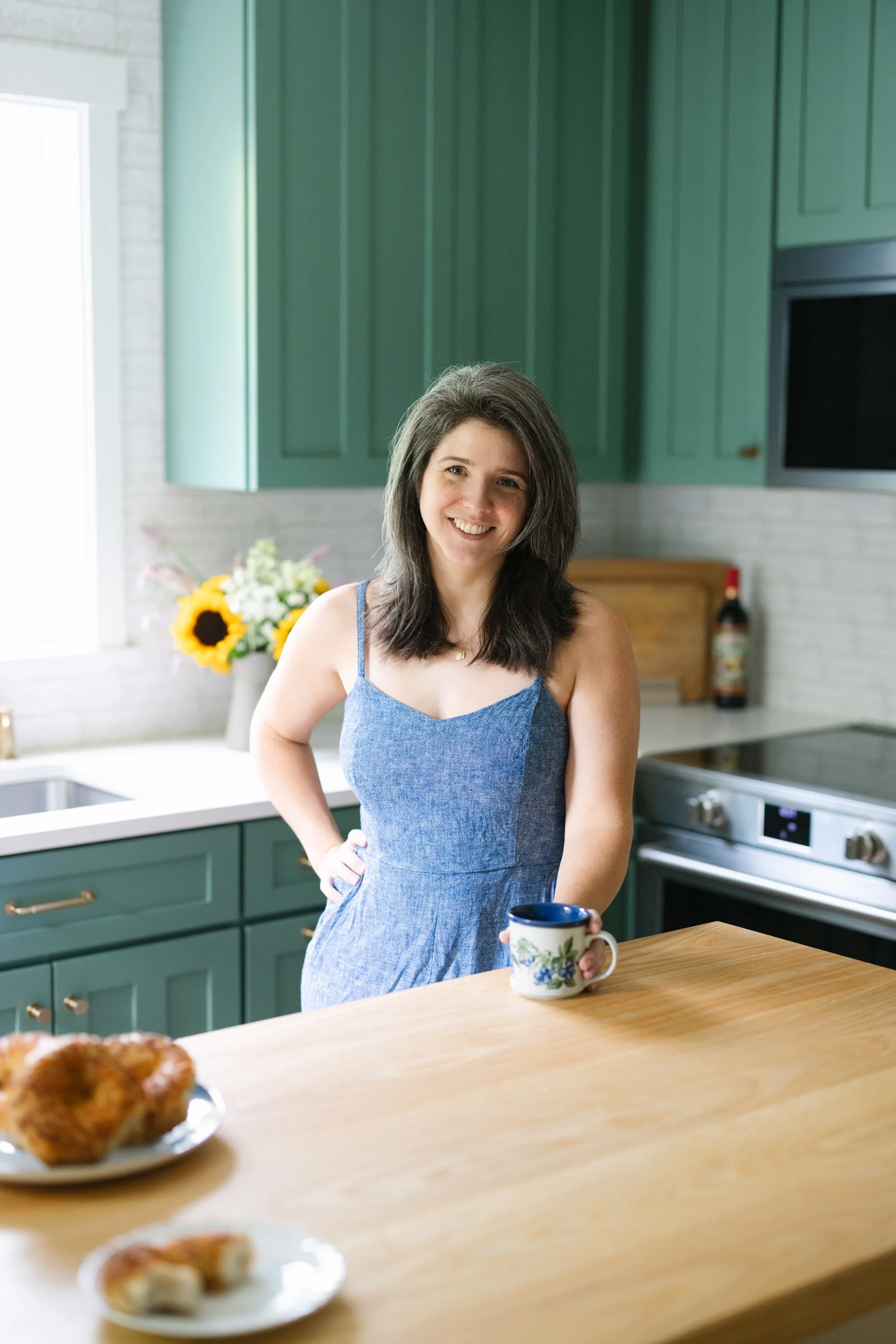 A woman with shoulder-length brown hair wearing a blue sleeveless dress, smiling and holding a floral-patterned mug, standing in a kitchen with green cabinets and a window in the background.