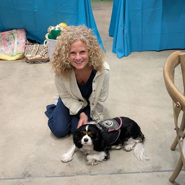 A woman with curly blonde hair kneeling on the floor next to a black and white Cavalier King Charles Spaniel, both in an indoor setting with blue curtains and luggage in the background.