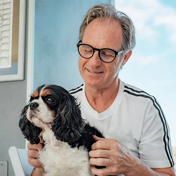 A smiling man with glasses gently holding a small black, white, and tan Cavalier King Charles Spaniel dog.