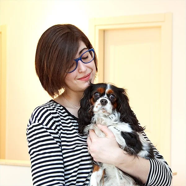 A woman with short brown hair, wearing blue glasses and a black-and-white striped shirt, holding a small black, white, and brown Cavalier King Charles Spaniel puppy indoors.