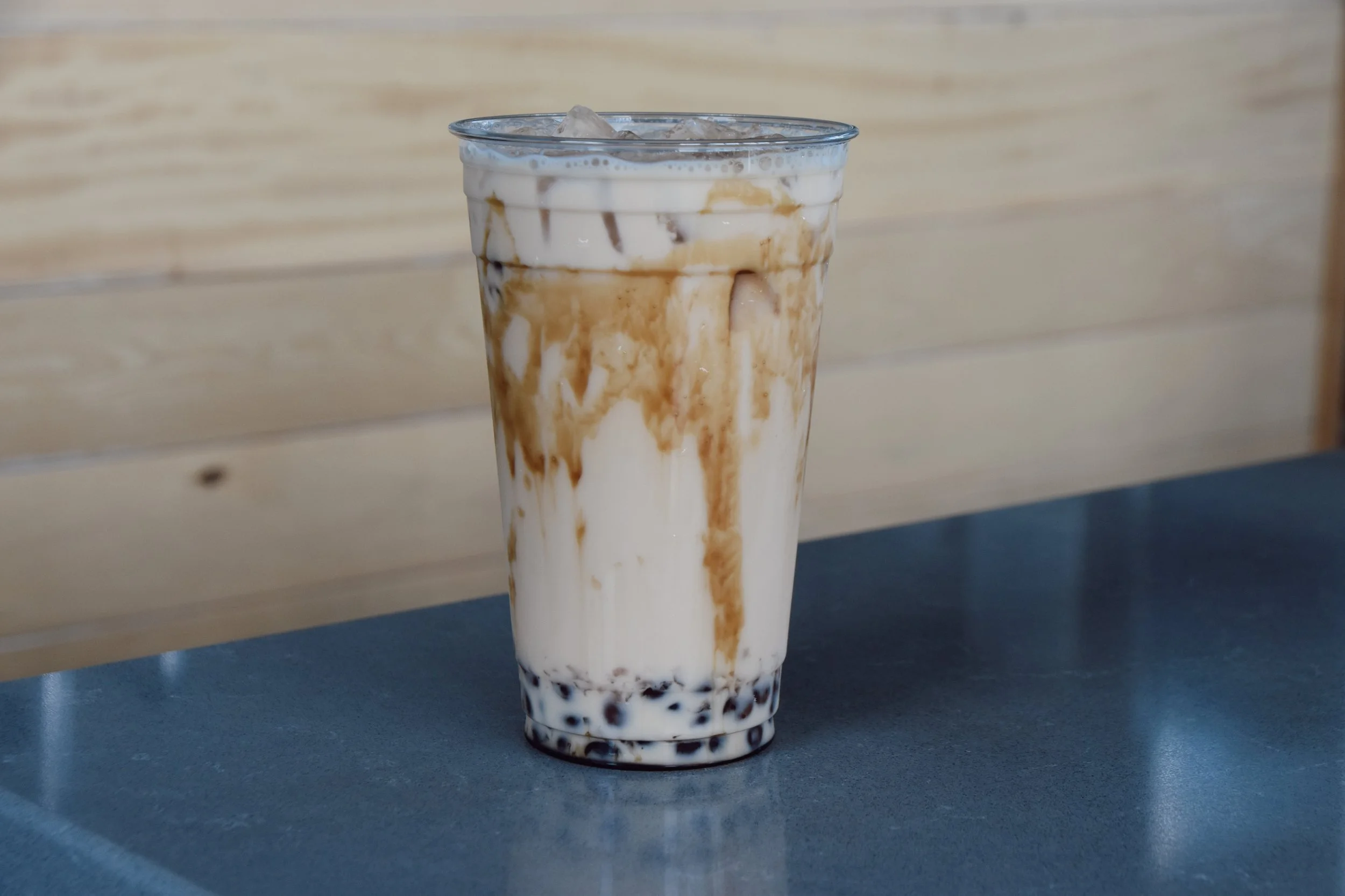 A tall glass of iced coffee with layers of milk and coffee, topped with ice cubes and caramel syrup, on a dark blue table with a light wood background.