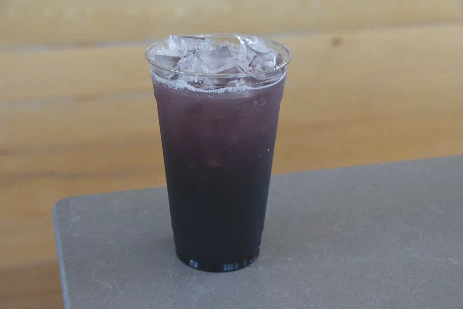A clear glass filled with a dark purple beverage and ice cubes on top, placed on a gray surface with a wooden background.