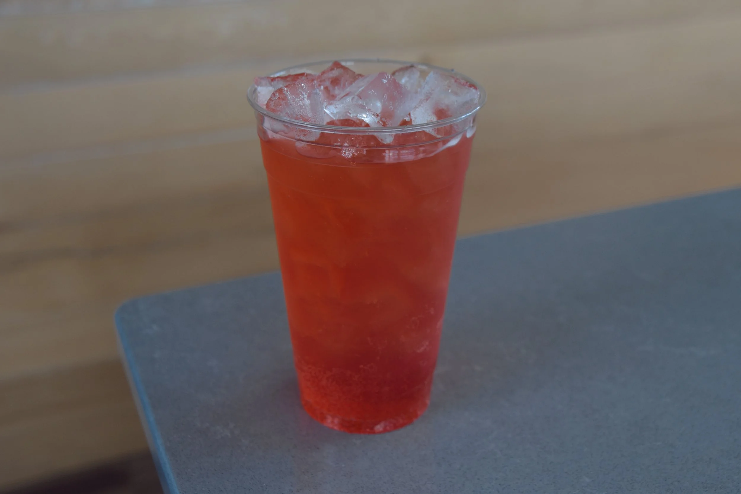 A clear plastic cup filled with a red-colored beverage and ice cubes resting on top.