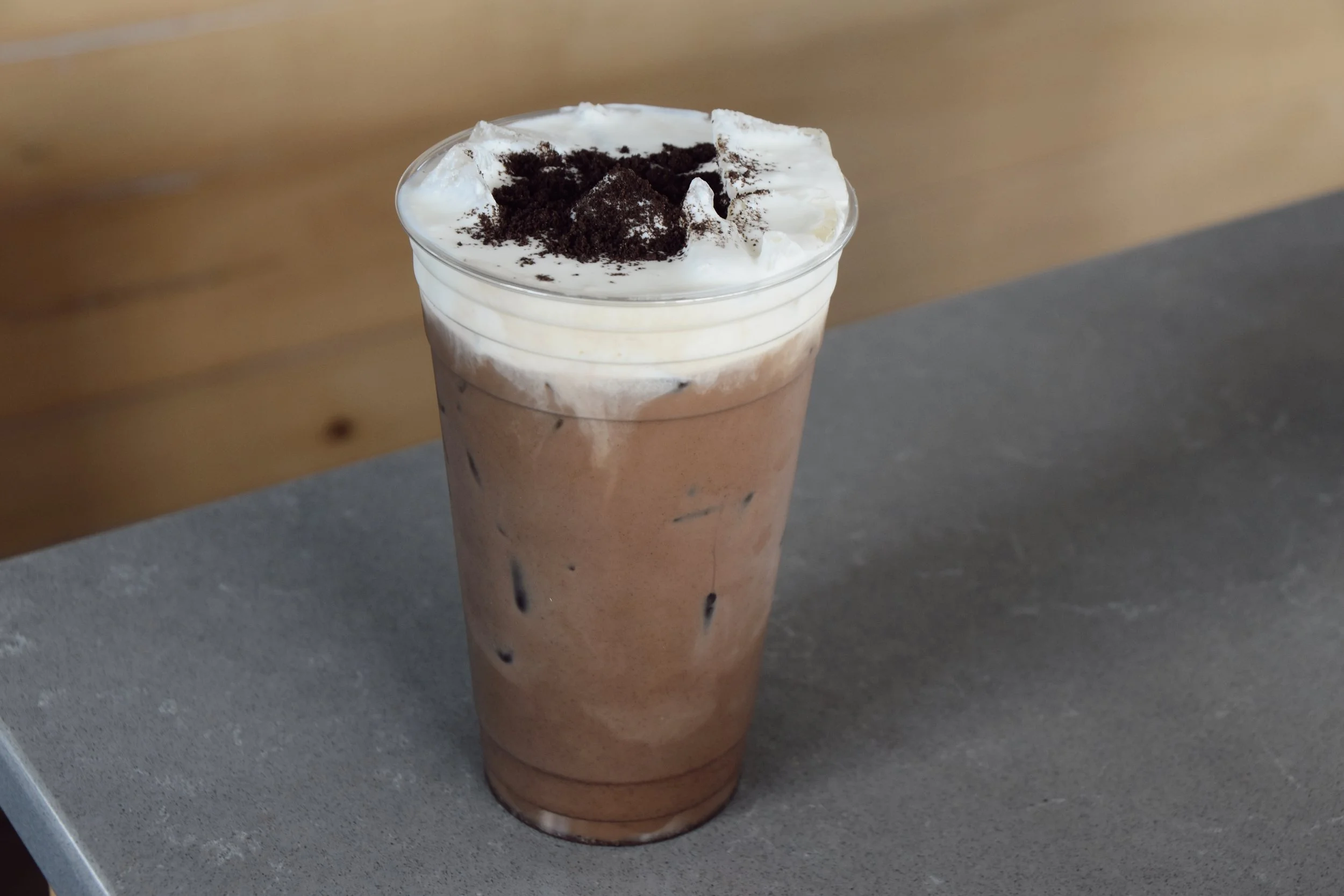 A tall glass of chocolate iced coffee topped with whipped cream and crushed Oreo cookies, sitting on a gray surface.