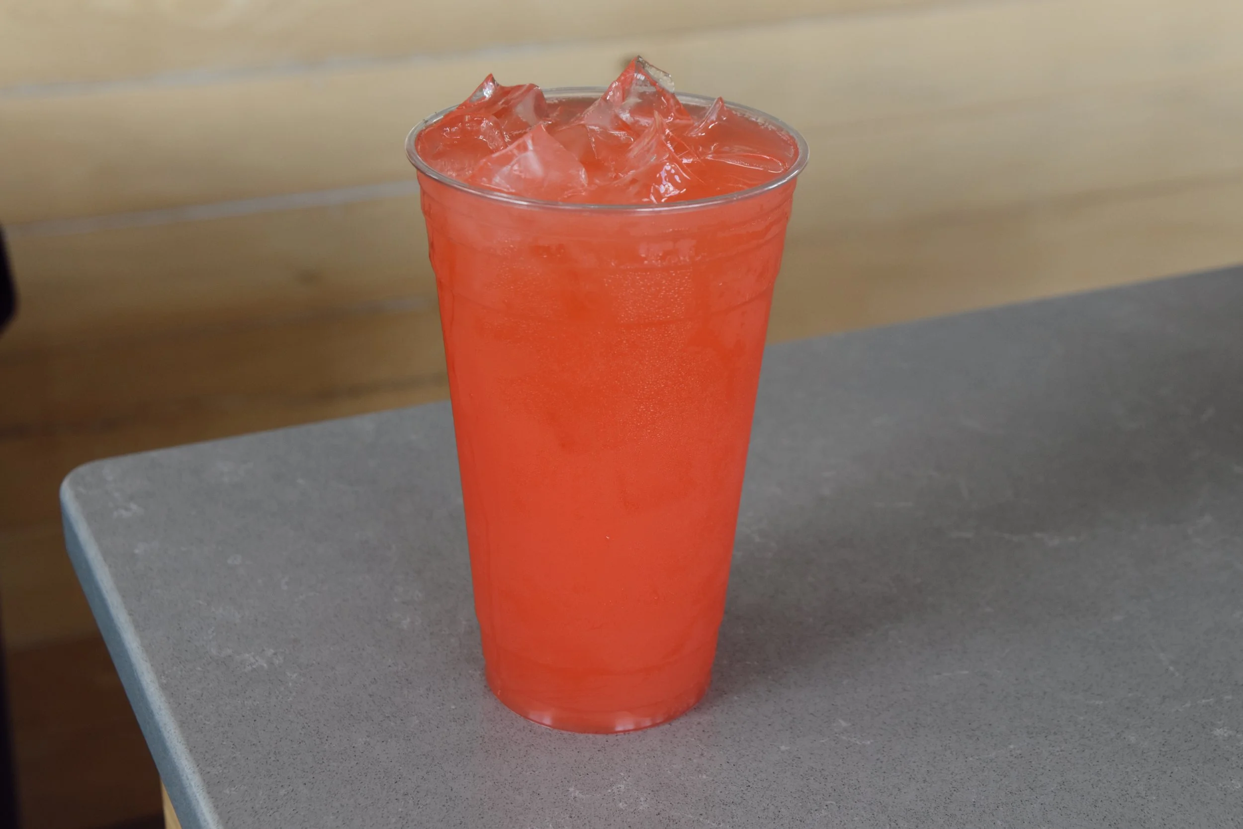 A clear plastic cup filled with orange-colored beverage and ice cubes, placed on a grey surface with a wooden background.