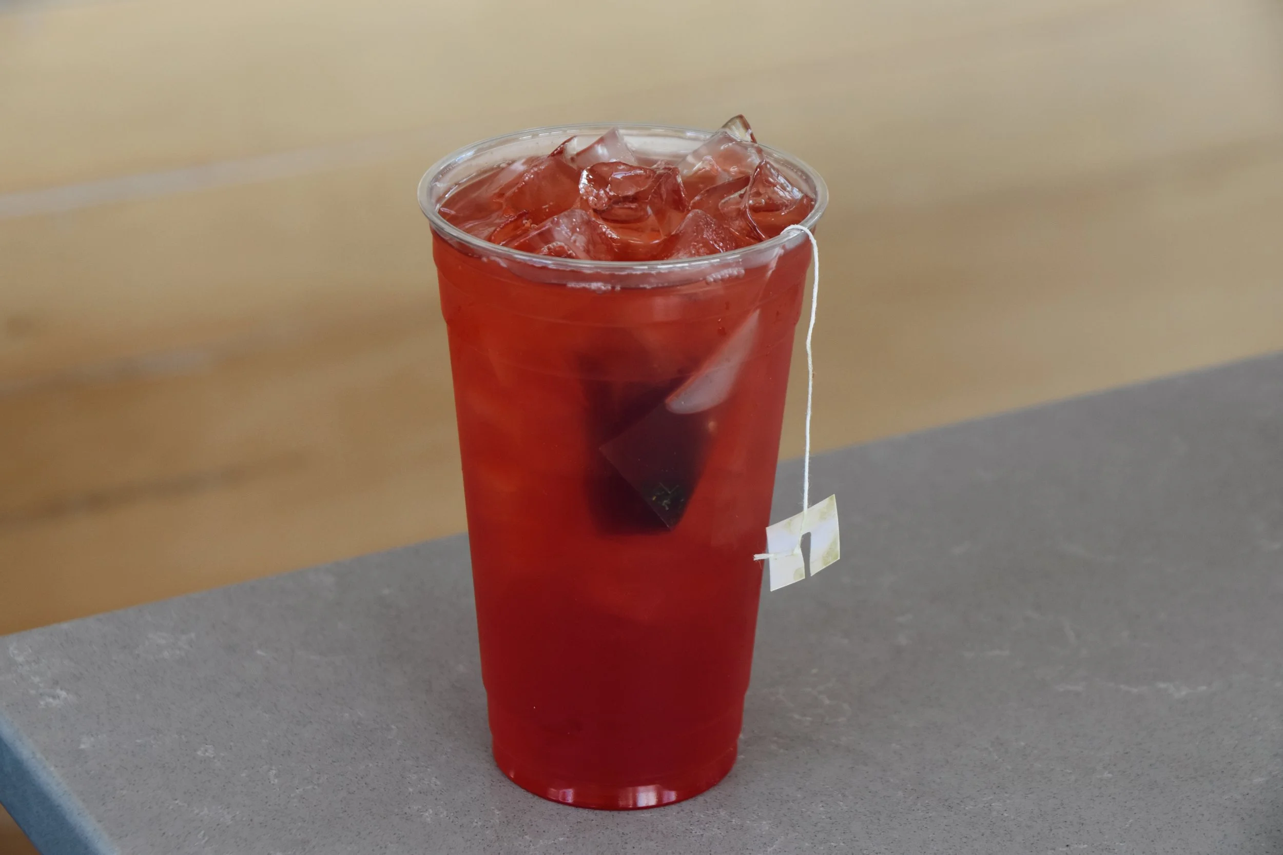 A clear plastic cup filled with ice cubes and red-colored beverage, placed on a gray surface with a plain light-colored background.