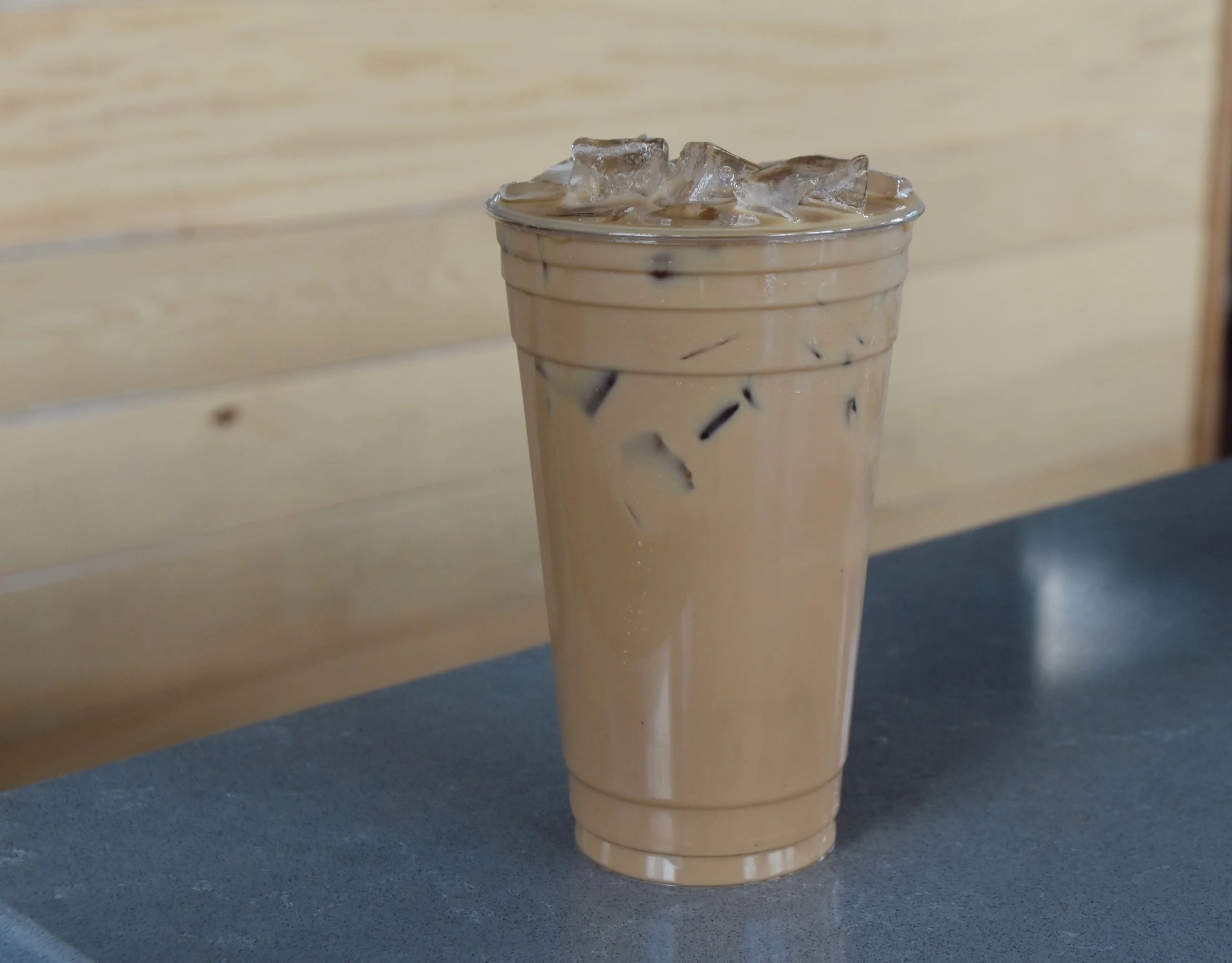A clear plastic cup filled with iced coffee or tea, topped with ice cubes, sitting on a dark surface in front of a light wood background.