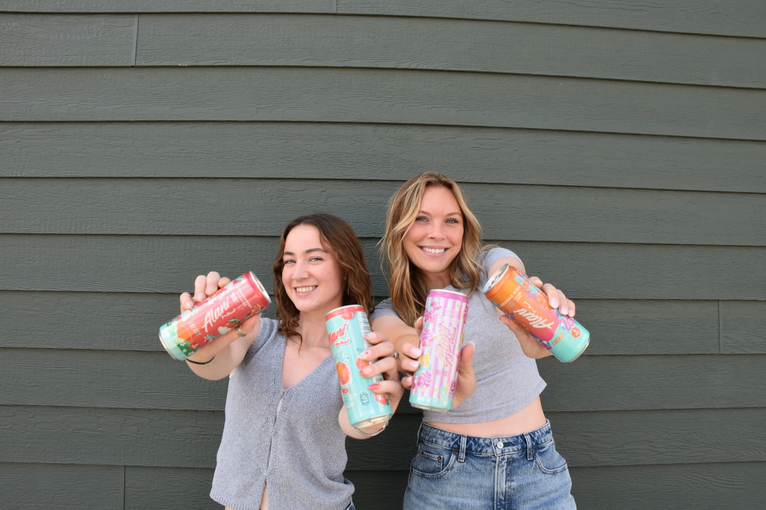 Two smiling young women holding colorful cans of energy drinks in front of a green wooden wall.