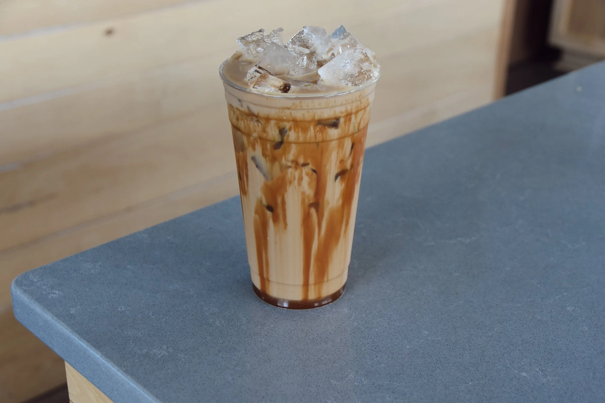 A tall glass of iced coffee with caramel drizzle and ice cubes on top, placed on a gray table.