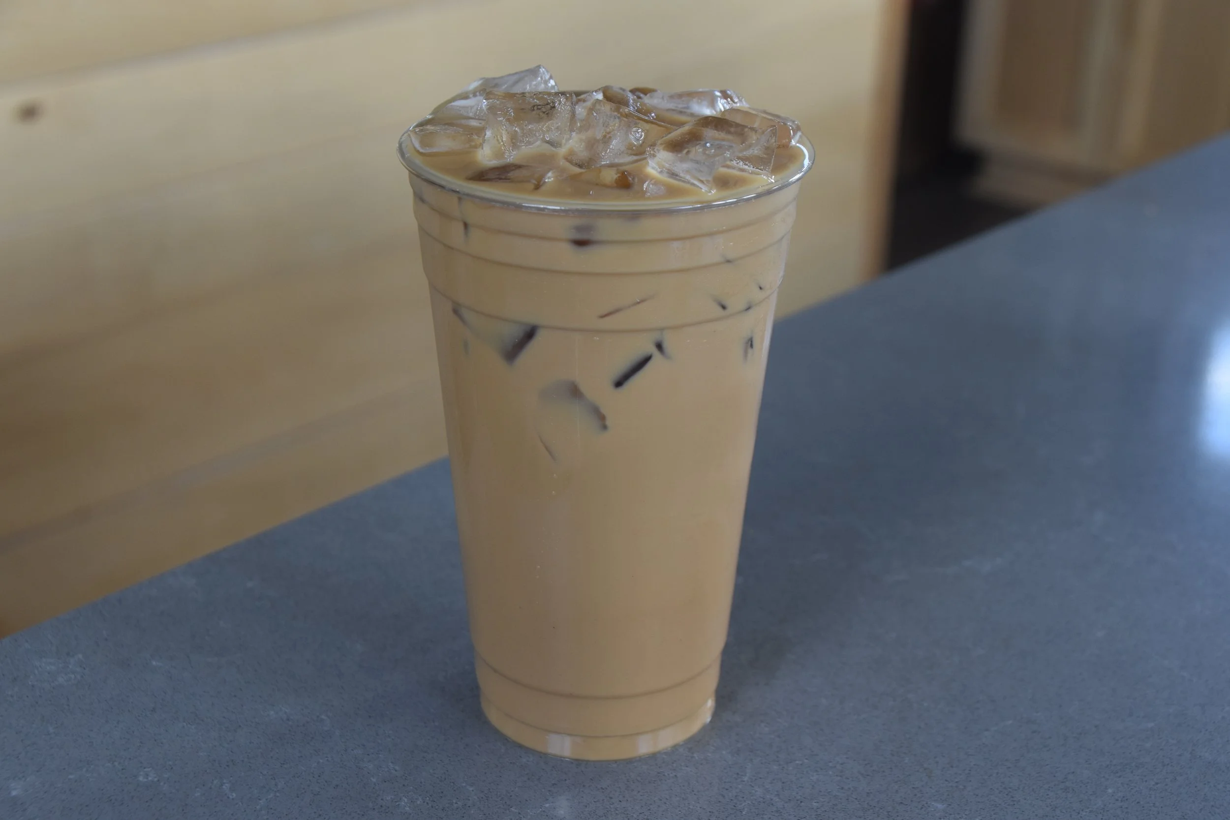 A tall clear plastic cup filled with iced coffee or iced latte, with ice cubes floating on top, placed on a dark countertop with a light-colored wall in the background.