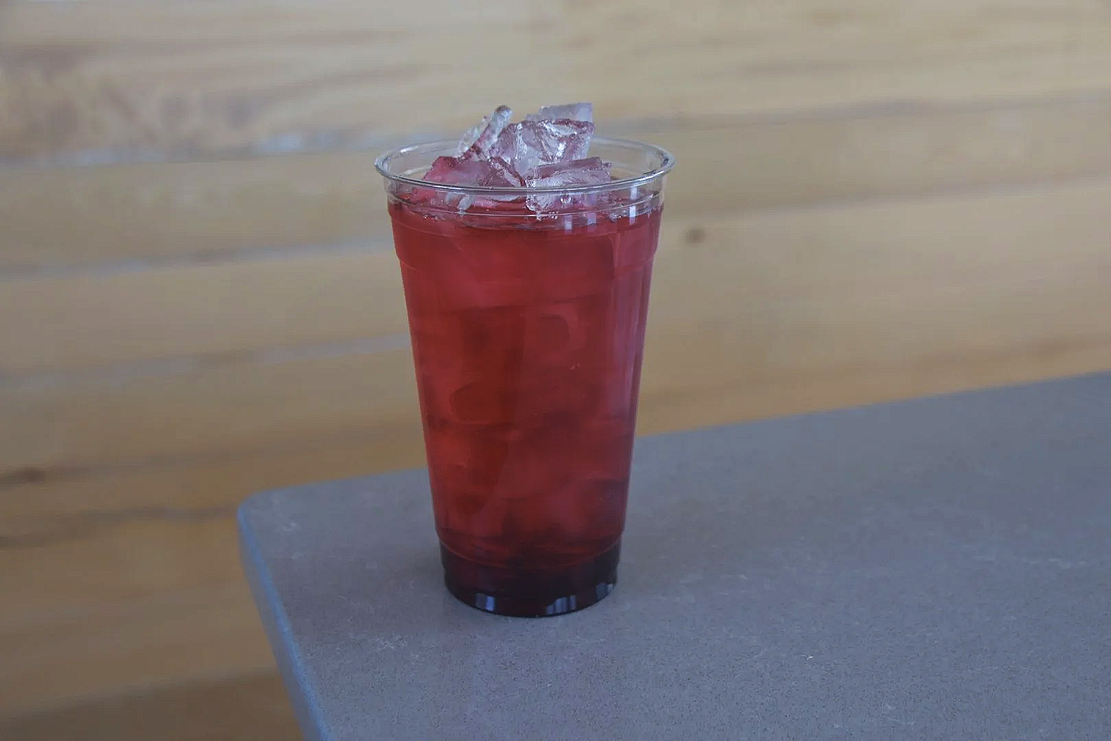 A clear plastic cup filled with a red beverage and ice cubes, placed on a gray surface with a wooden background.