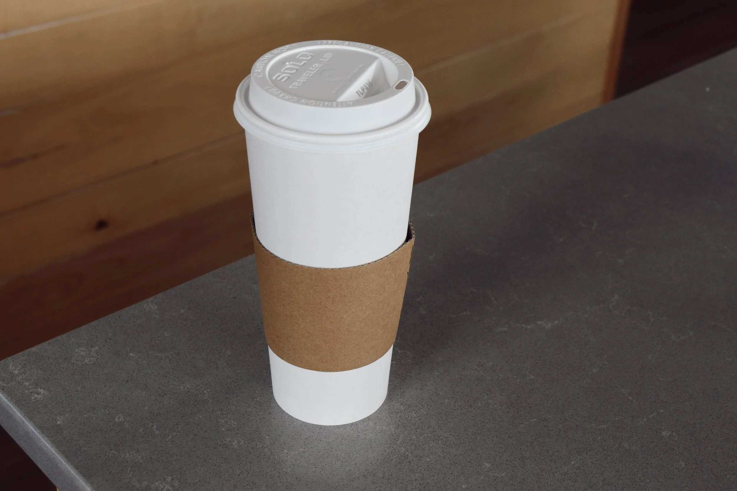 A white disposable coffee cup with a brown cardboard sleeve and a white plastic lid, sitting on a gray surface with a wooden wall in the background.