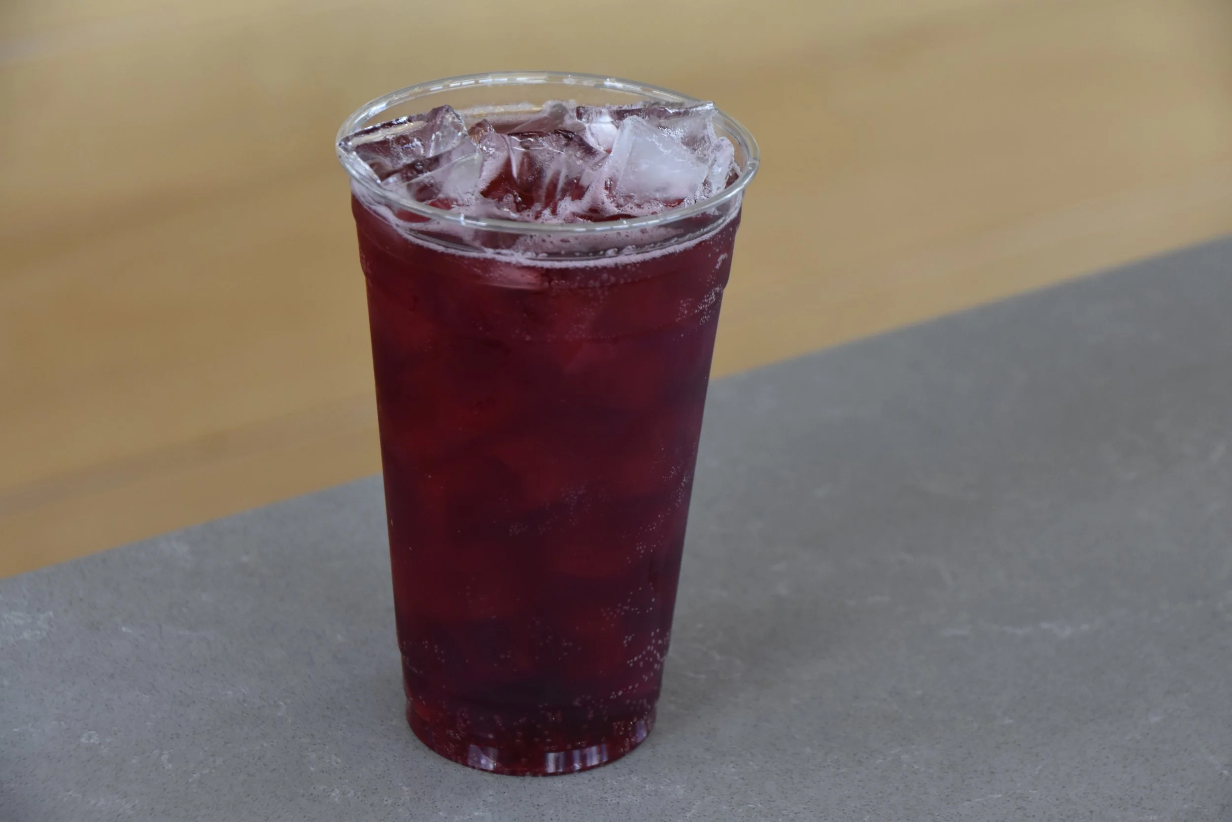 A tall clear glass filled with dark purple beverage and ice cubes, placed on a gray surface with a yellow and gray background.