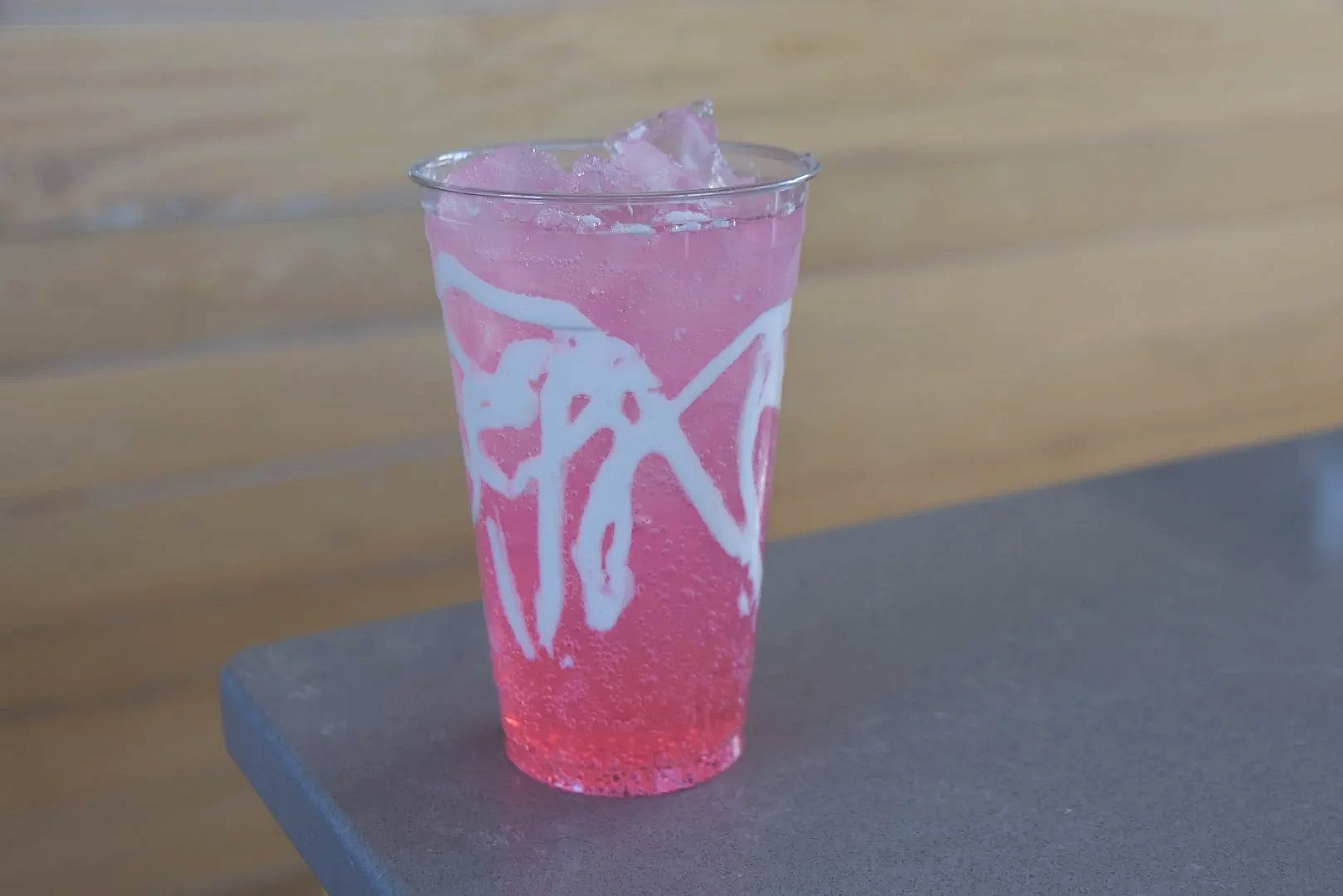A clear glass filled with pink iced beverage with white decorative drizzle, placed on a dark gray surface, with a wooden wall background.