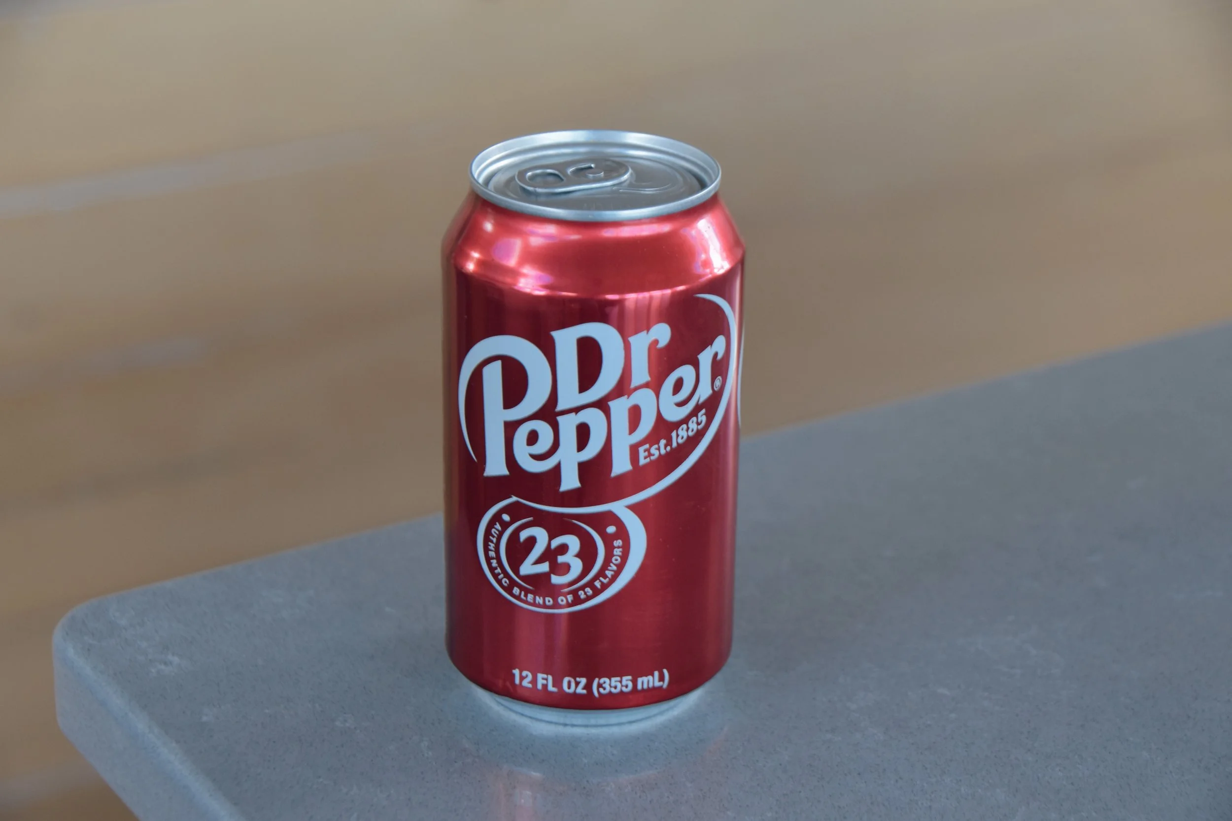 A 12-ounce can of Dr. Pepper with a red metallic finish and white and blue branding, placed on a gray surface with a beige and gray background.