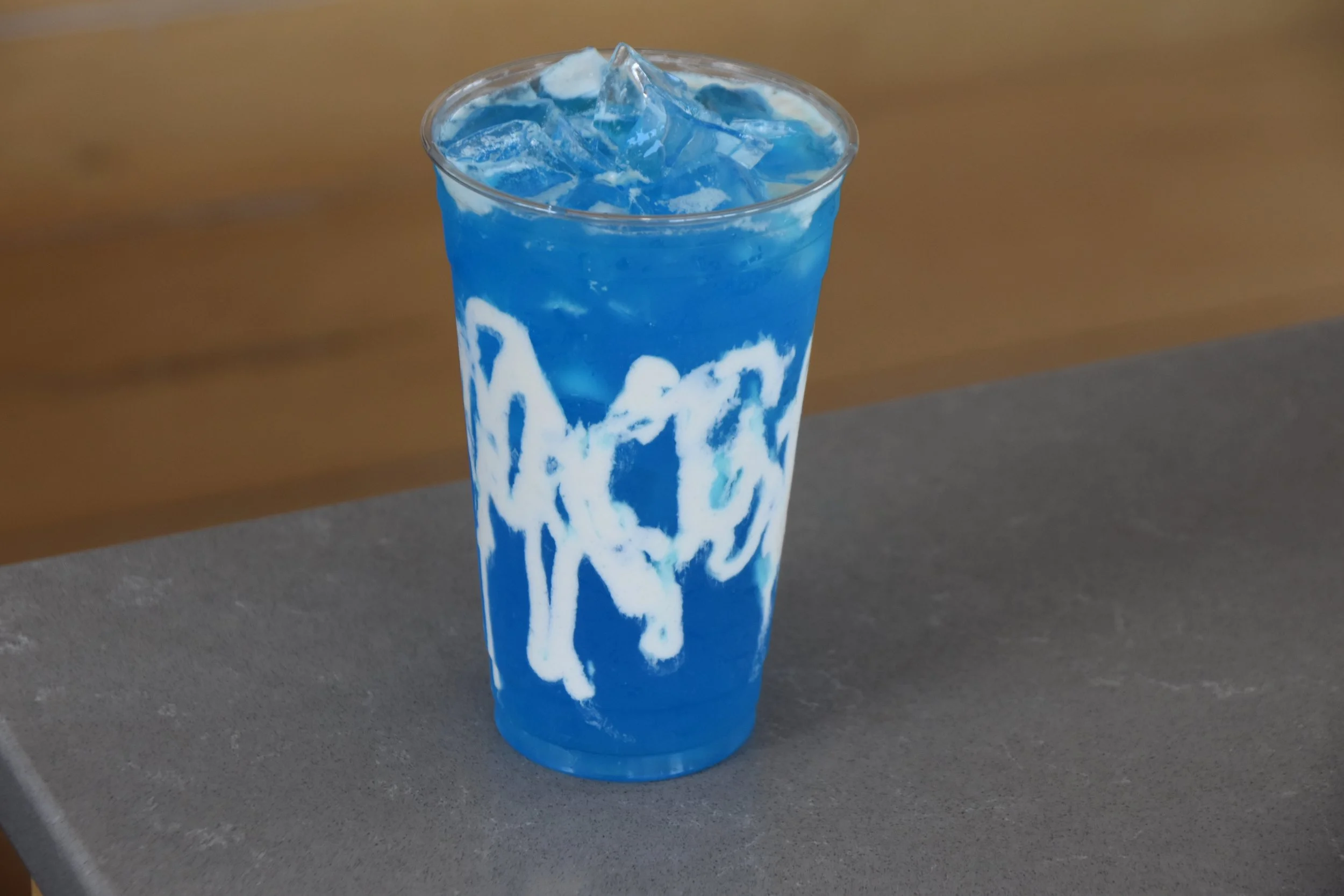 A tall glass filled with bright blue frozen slush topped with ice cubes, served on a gray table with a wooden background.