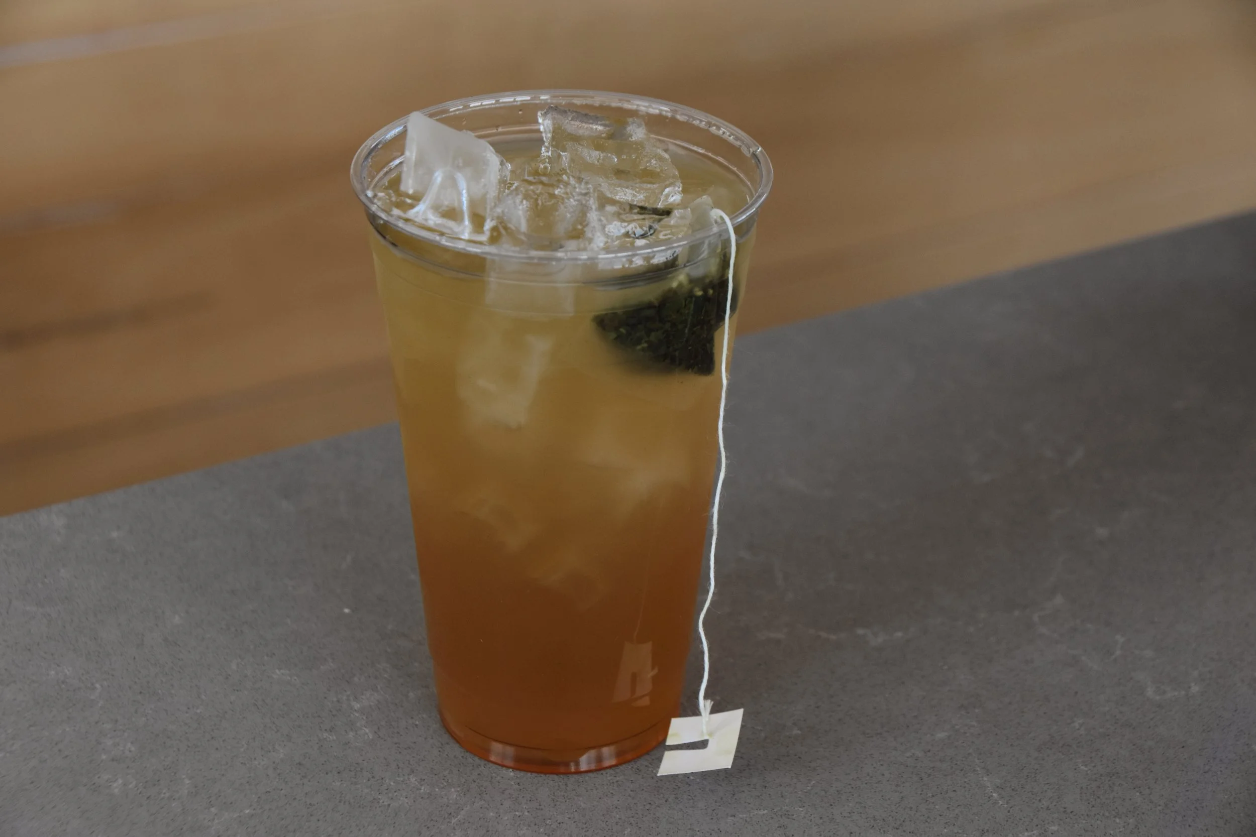 A tall plastic cup filled with iced tea, with ice cubes and a tea bag inside, sitting on a gray surface with a wooden background.