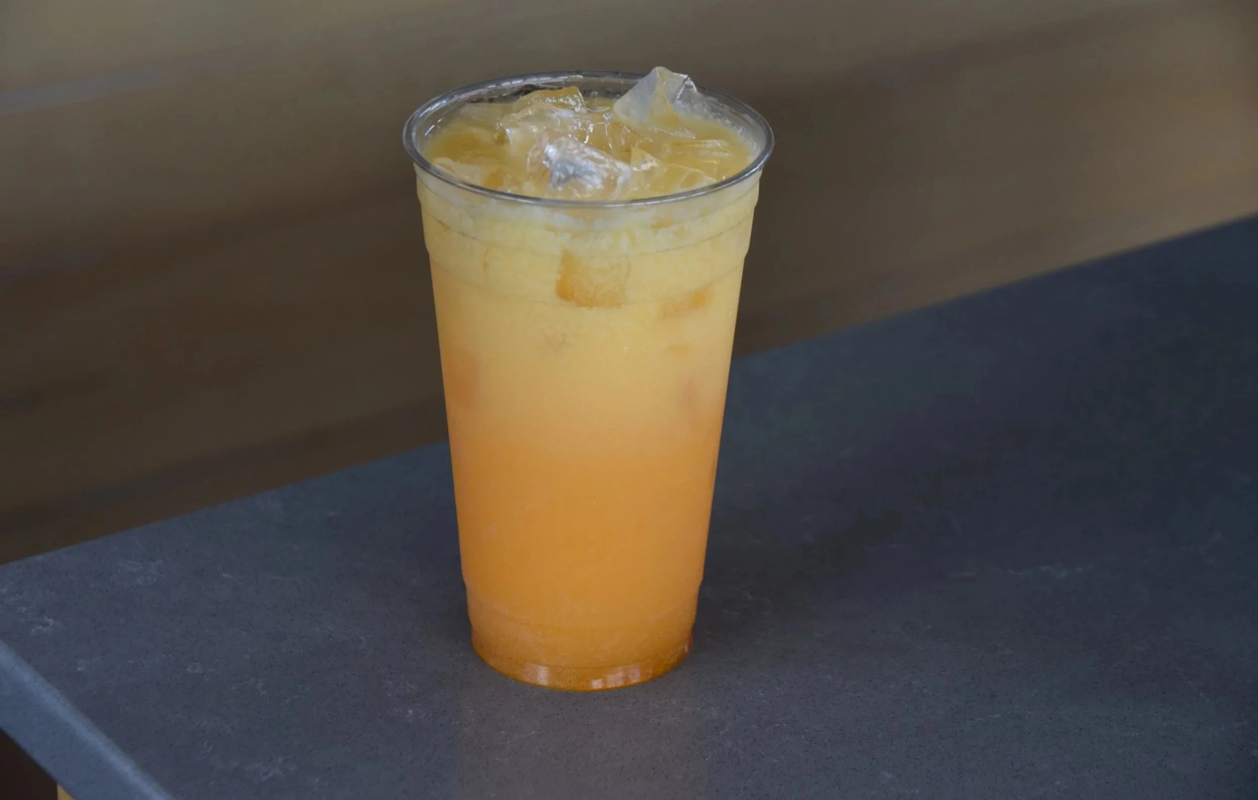 A tall transparent glass filled with a bright orange beverage, ice cubes floating on top, placed on a dark surface.