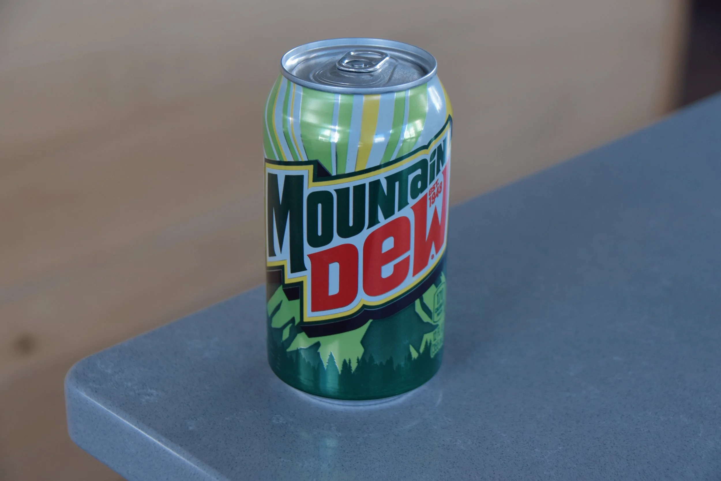 A can of Mountain Dew, a green soda can with colorful design, sitting on a light gray table with a rounded edge.