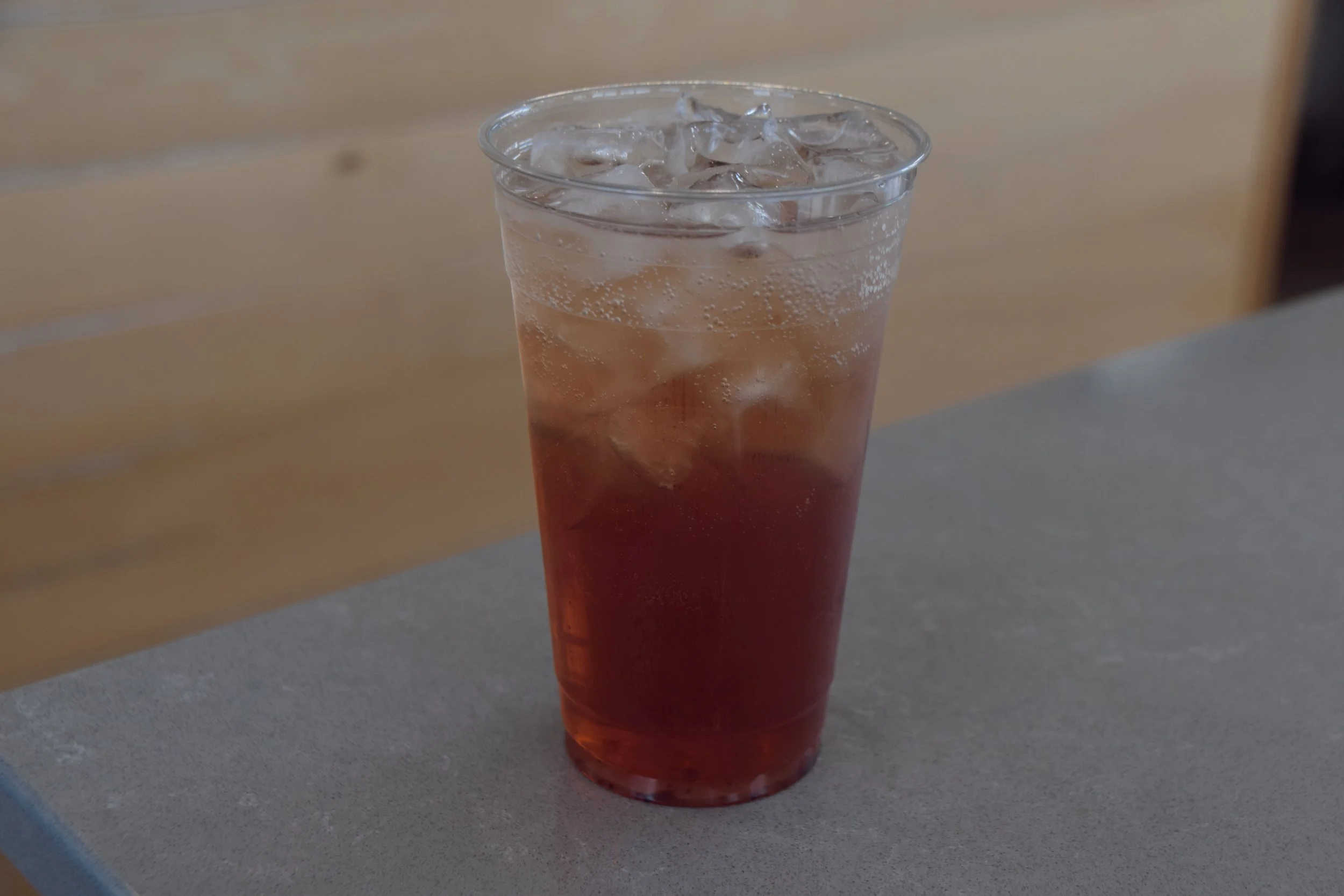 A clear plastic cup filled with a reddish-brown soda and ice cubes, placed on a gray surface with a wooden background.