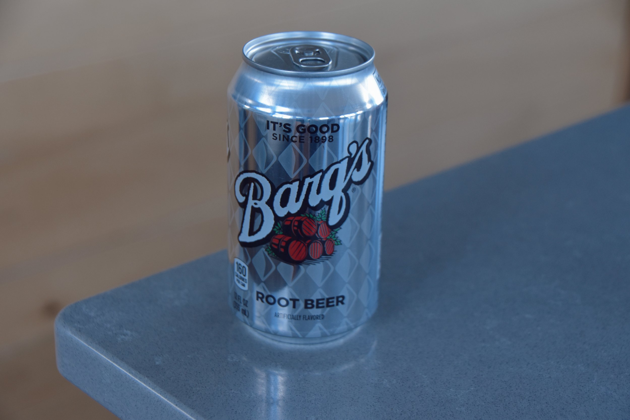 A can of root beer with the label 'Barq's' sits on a gray surface. The can is silver with a diamond pattern and red root beer graphics, and features the text 'It's good since 1898'.