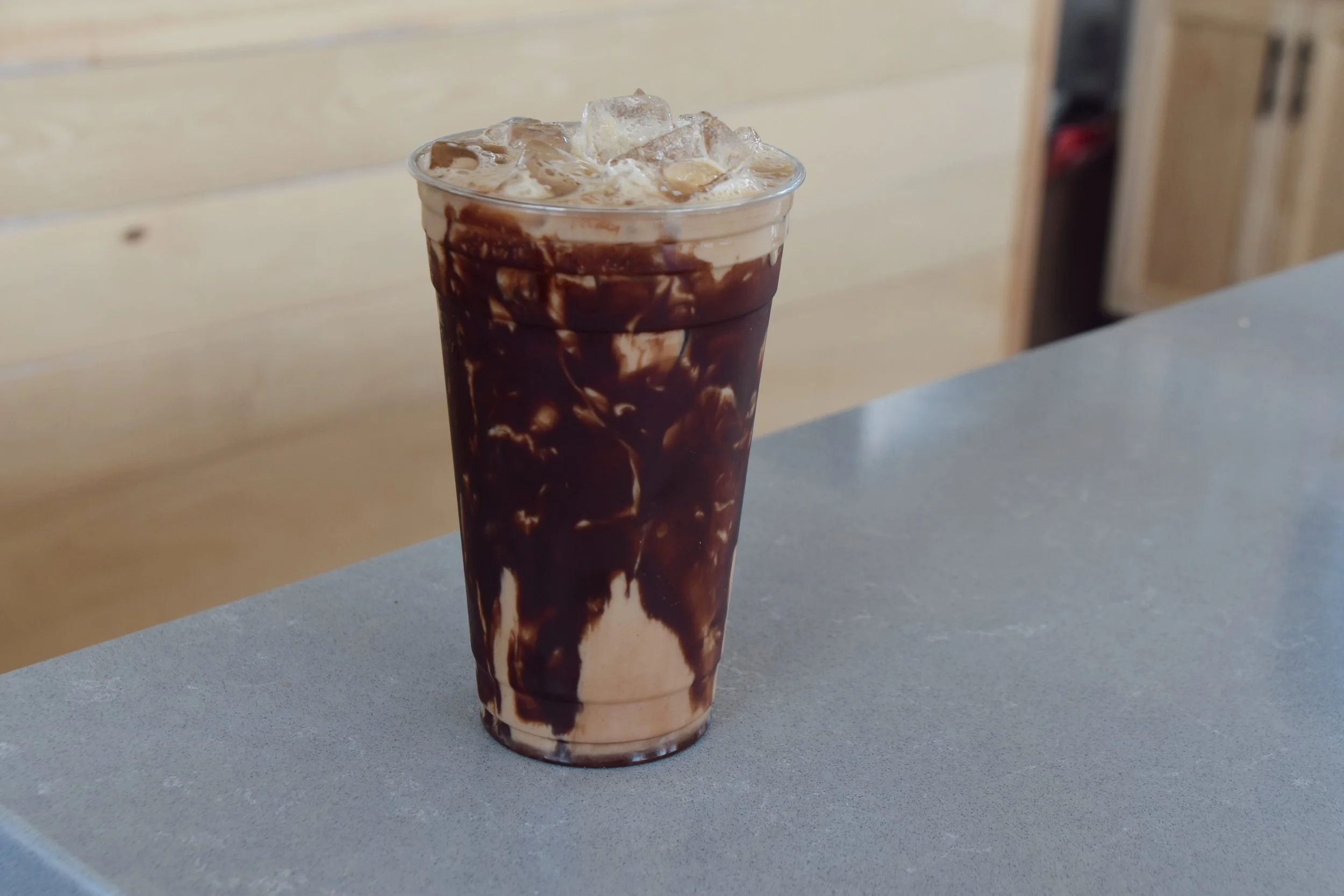 Tall glass of iced coffee with chocolate syrup and ice cubes on a gray table, with a light-colored wooden wall in the background.