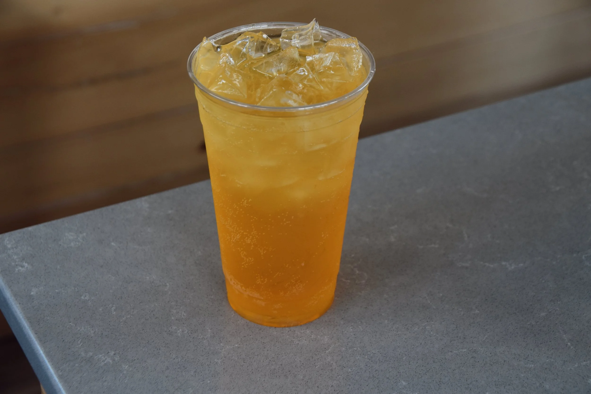 A clear plastic cup filled with orange-colored iced beverage topped with ice cubes, placed on a gray surface with a wooden background.