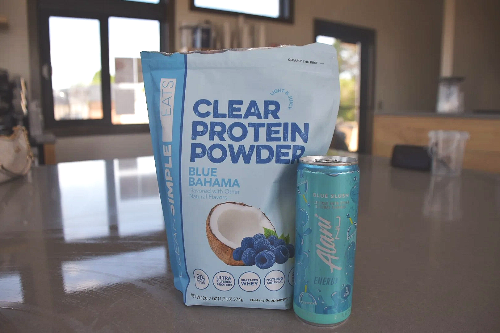 A bag of SlimPleasure Clear Protein Powder in Blue Bahama flavor and a blue energy drink can placed on a gray table in a room with windows and furniture in the background.