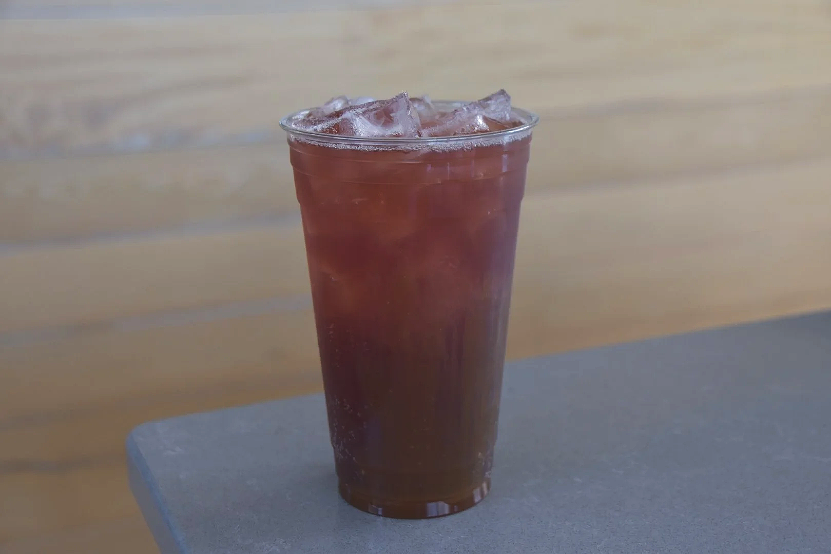 A glass filled with a dark purple cold beverage and ice cubes, placed on a gray surface with a wooden wall background.