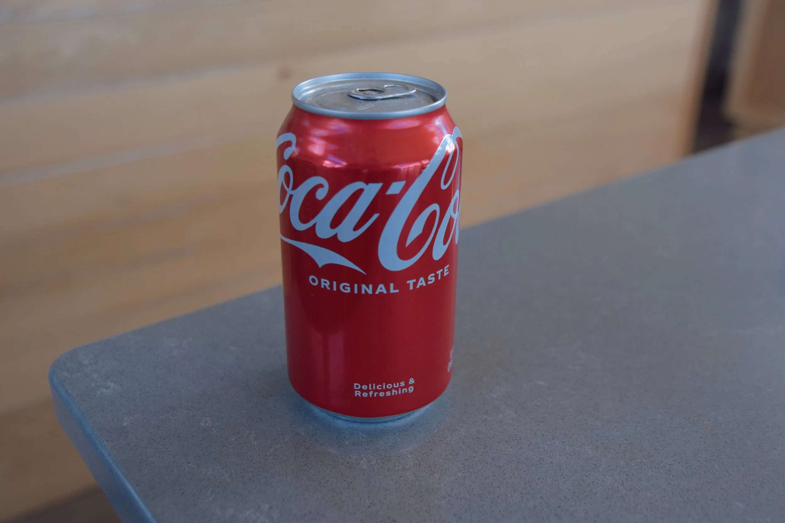 A red Coca-Cola can placed on a gray surface with a wooden background.