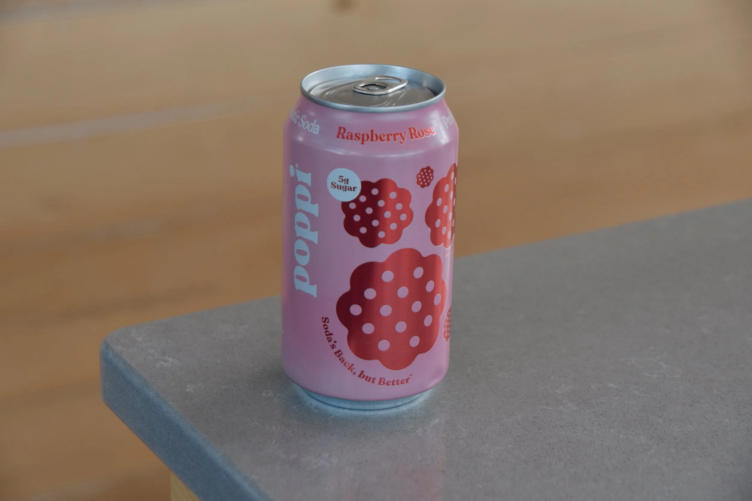 A pink aluminum soda can with raspberry flavor on a gray surface, against a wood background.
