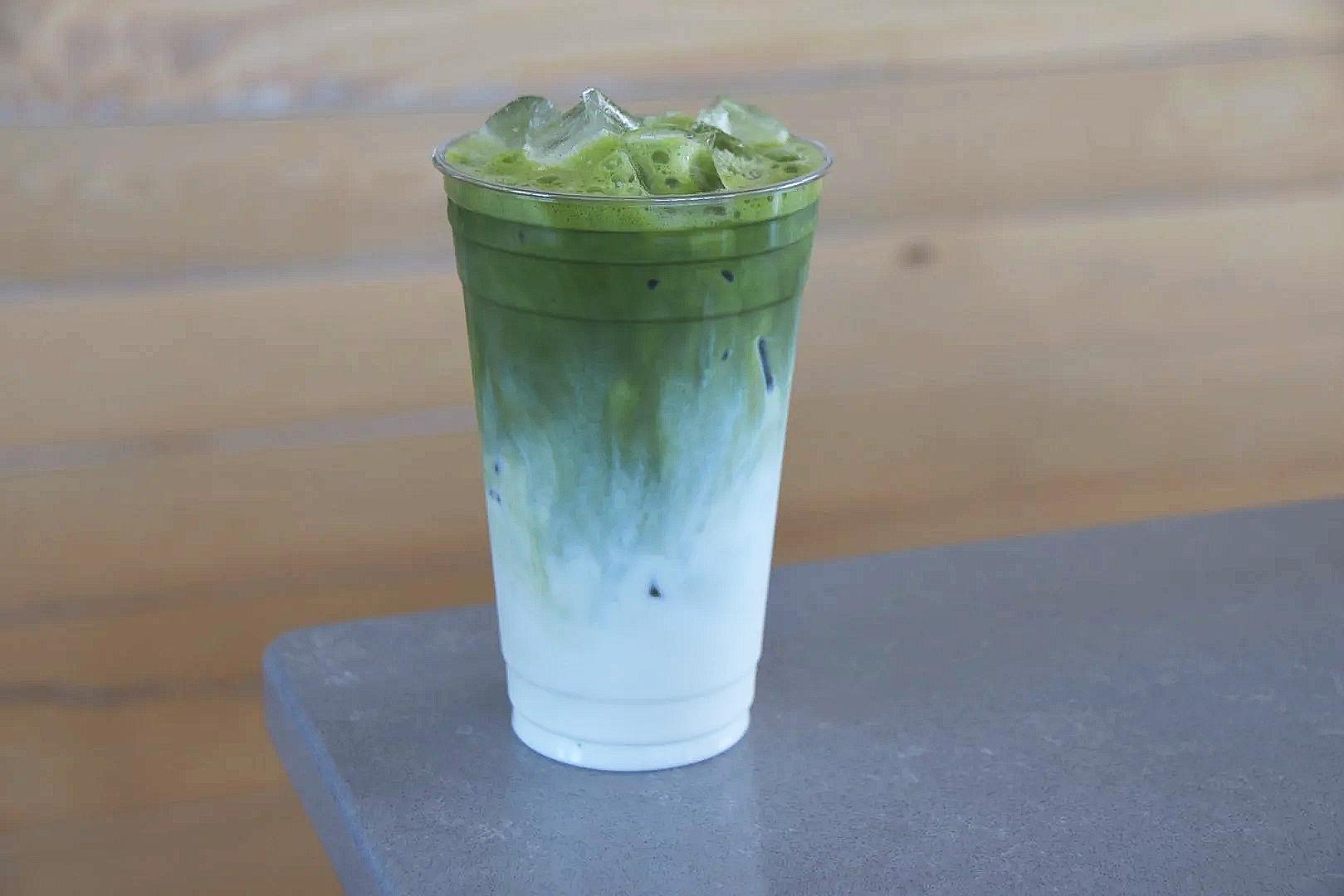 A plastic cup filled with iced matcha green tea latte, showing a layered effect with white milk at the bottom and vibrant green tea at the top, placed on a gray surface.