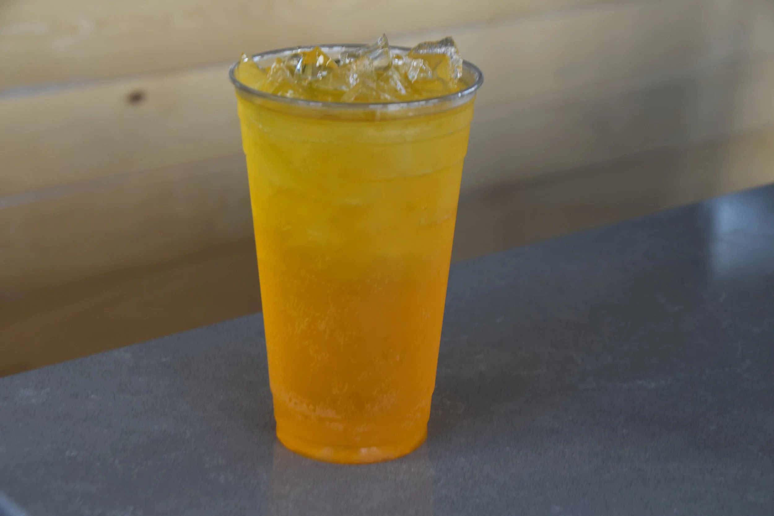 A clear plastic cup filled with yellow-orange beverage and ice cubes on a gray surface with a beige wooden background.