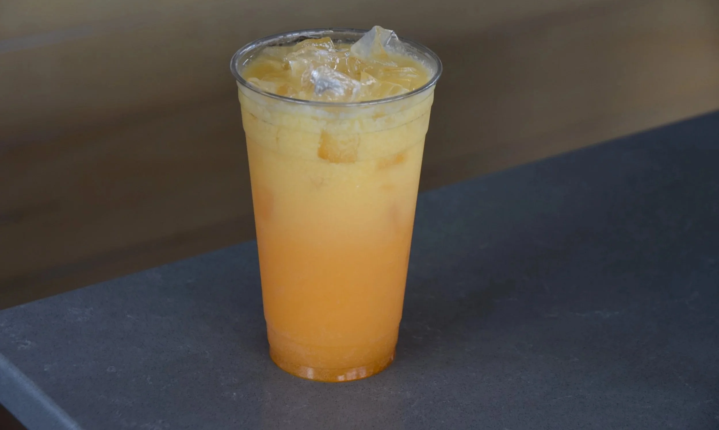 A clear plastic cup filled with orange-colored beverage and ice cubes, placed on a dark surface.