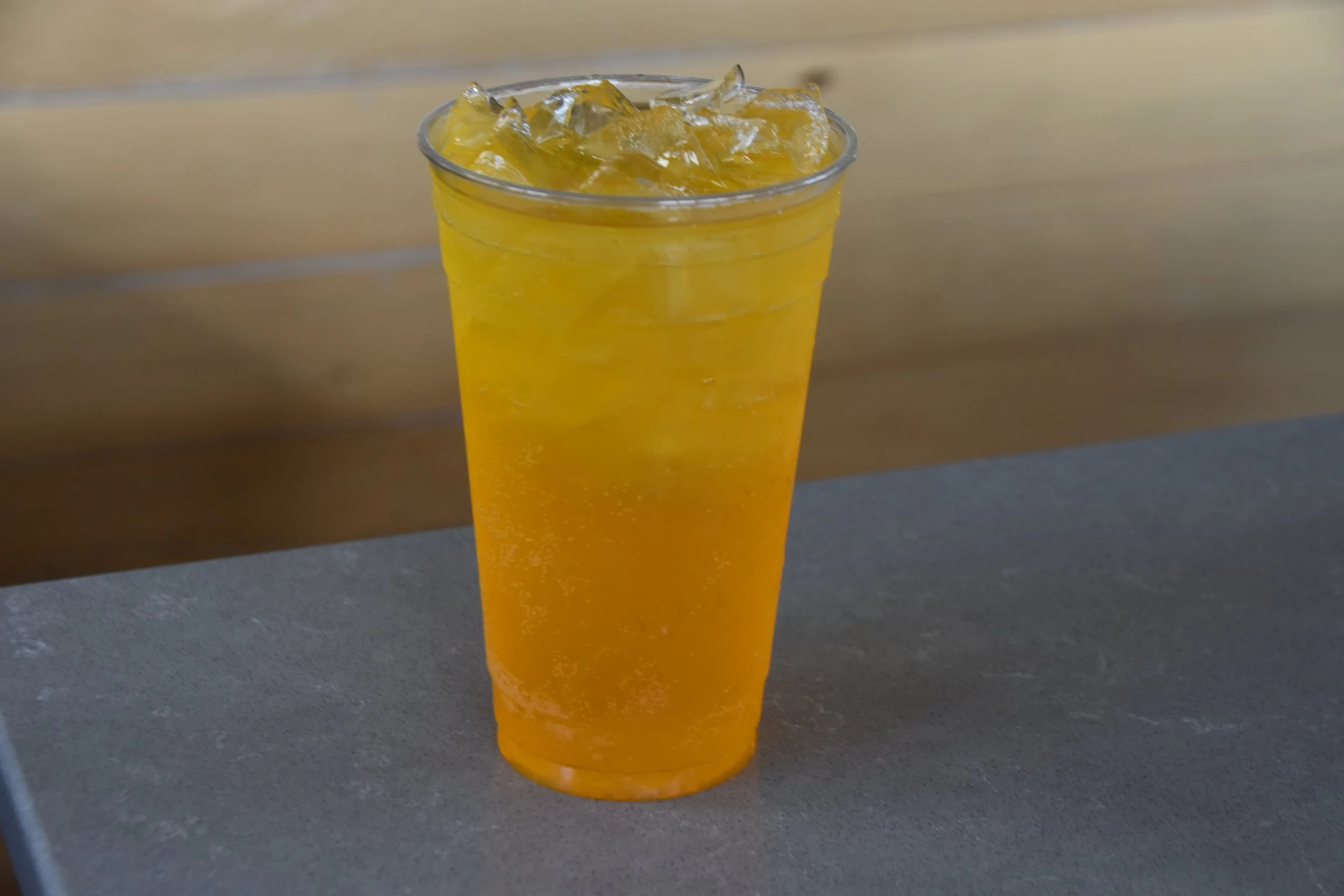 A clear plastic cup filled with yellow-orange beverage and ice cubes, sitting on a gray surface with a wooden background.