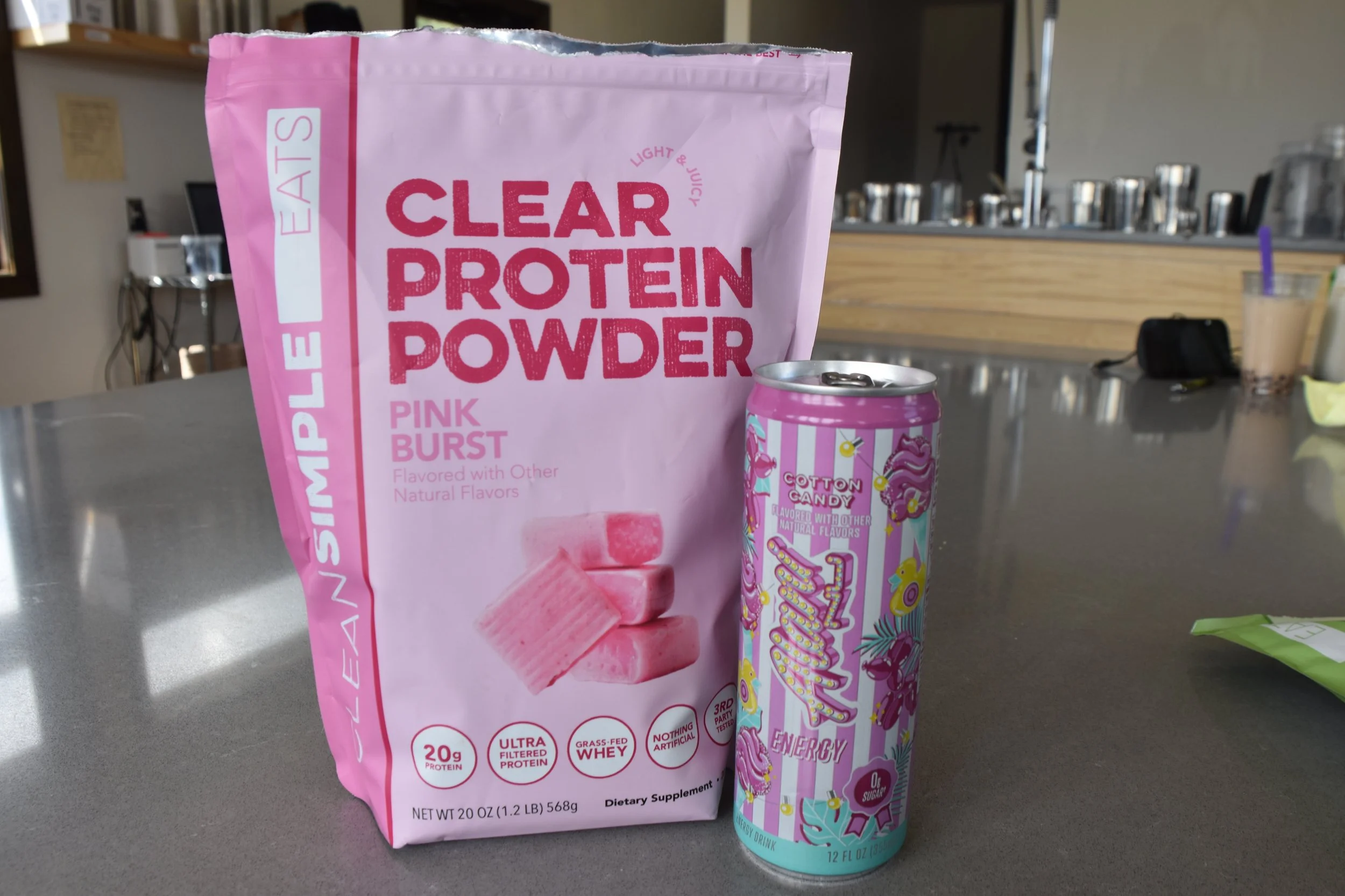 A pink bag of clear protein powder with flavor pink burst and a colorful energy drink can named Cotton Candy on a gray table.